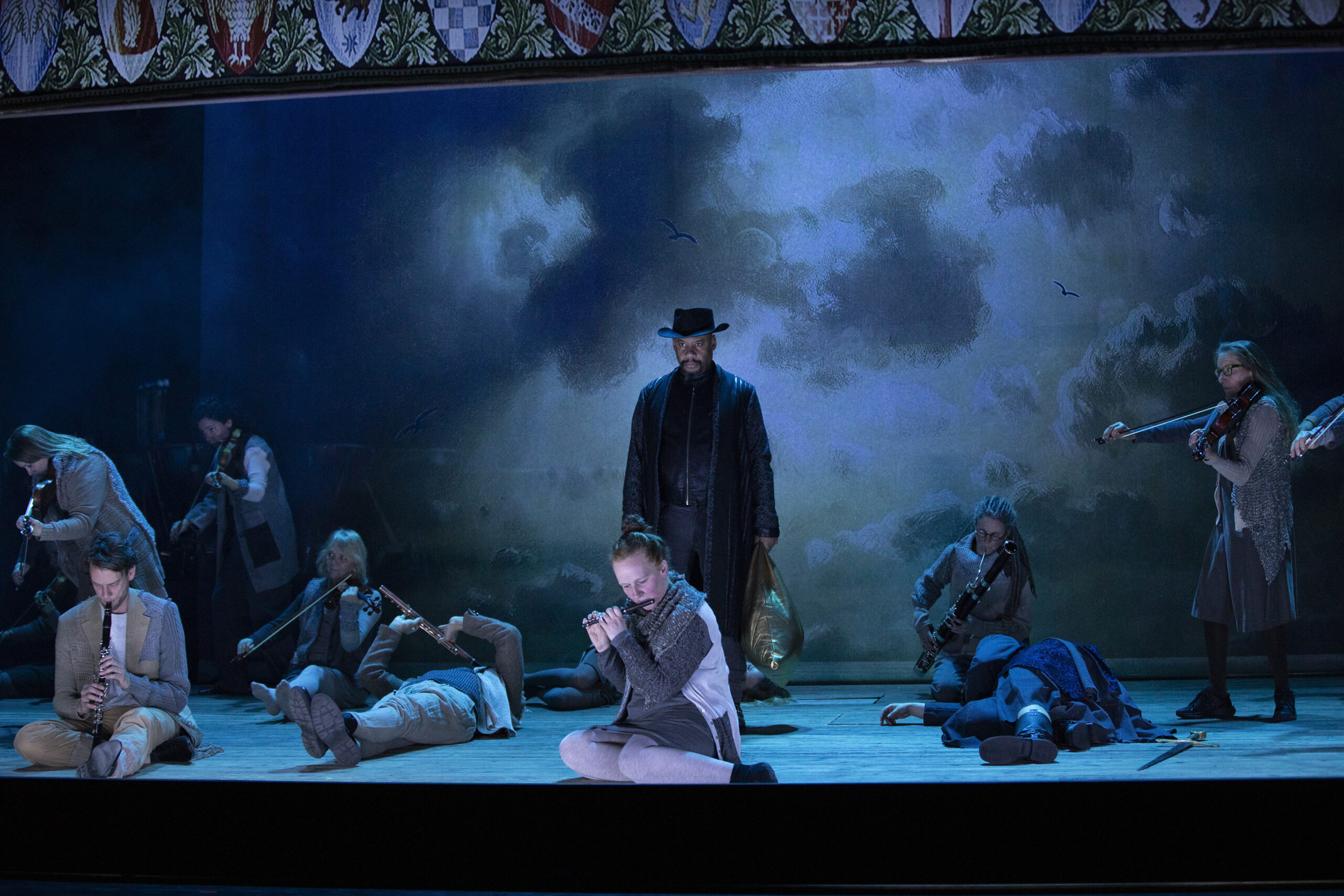 Op een donker podium liggen meerdere mensen met instrumenten op de grond terwijl een man in het midden staat met een zwarte hoed op