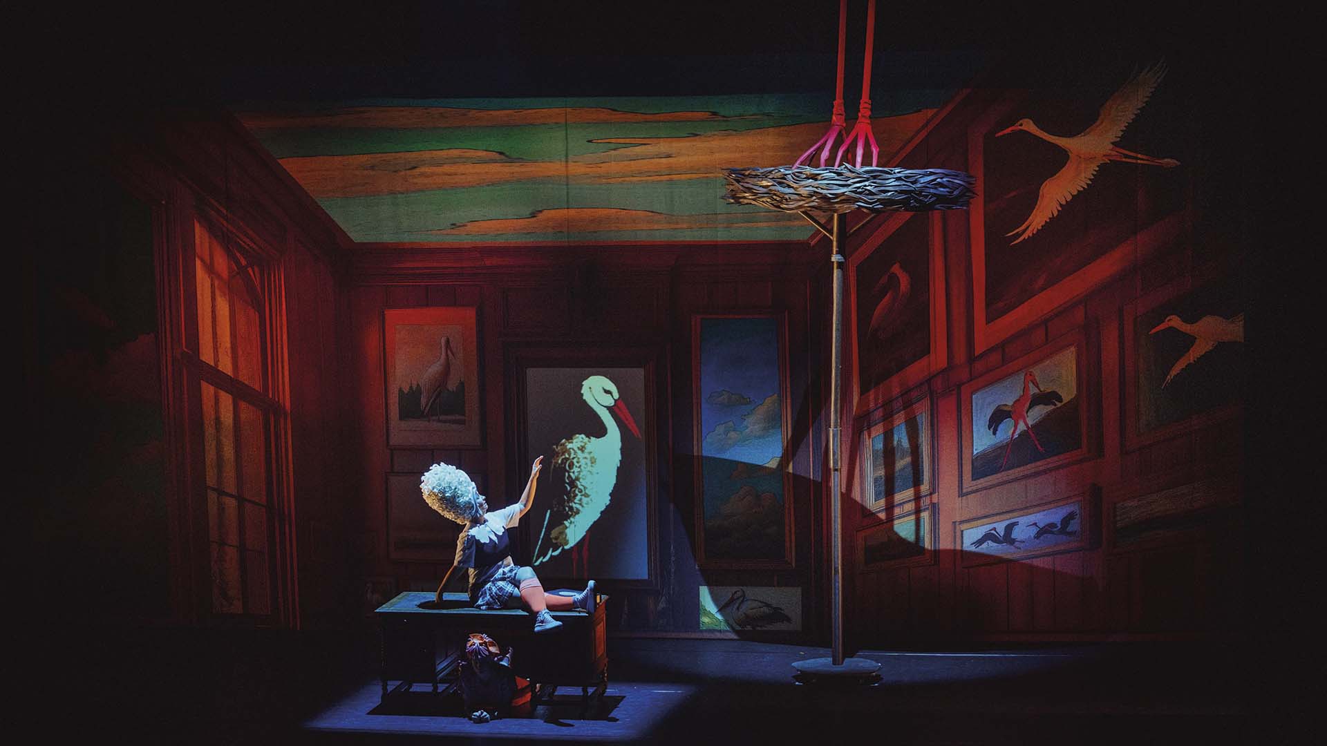 Theaterscène waarin een kind in witte pruik op tafel zit en wijst naar een projectie van een ooievaar, omringd door schilderijen in een klassiek ingerichte kamer.
