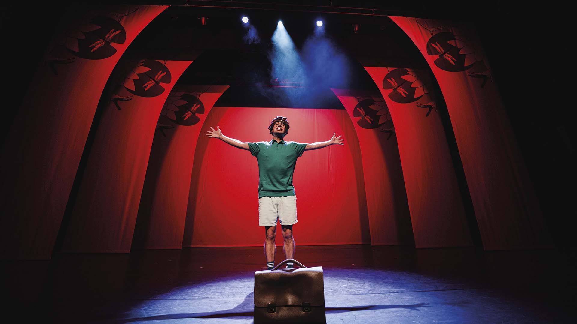Een acteur staat op een koffer midden op het toneel met zijn armen wijd, tegen een felrode achtergrond met decor van waterlelies en libellen.