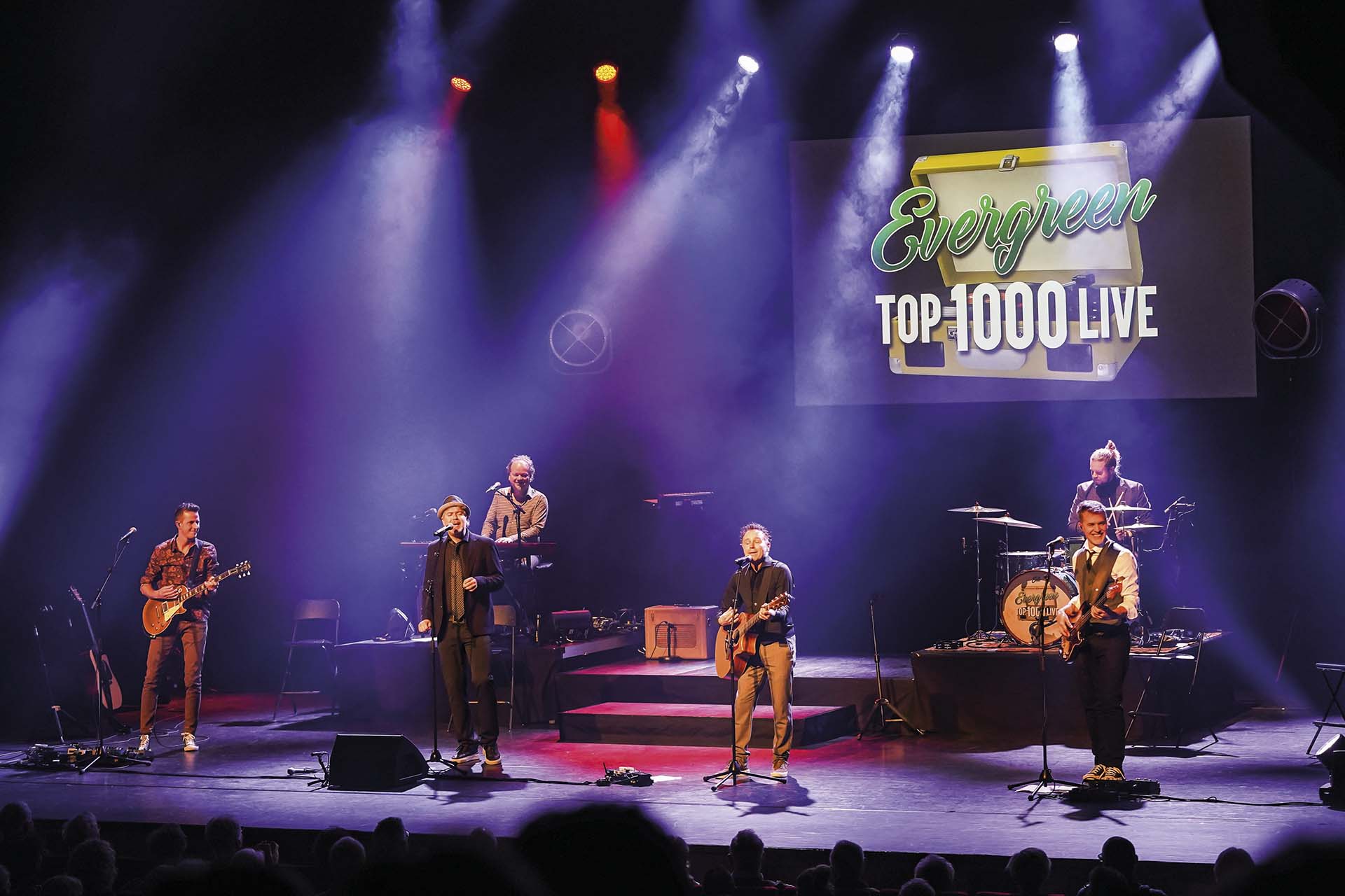 De volledige Evergreen Top 1000 Live band speelt op een verlicht podium met een projectie van het logo op de achtergrond.