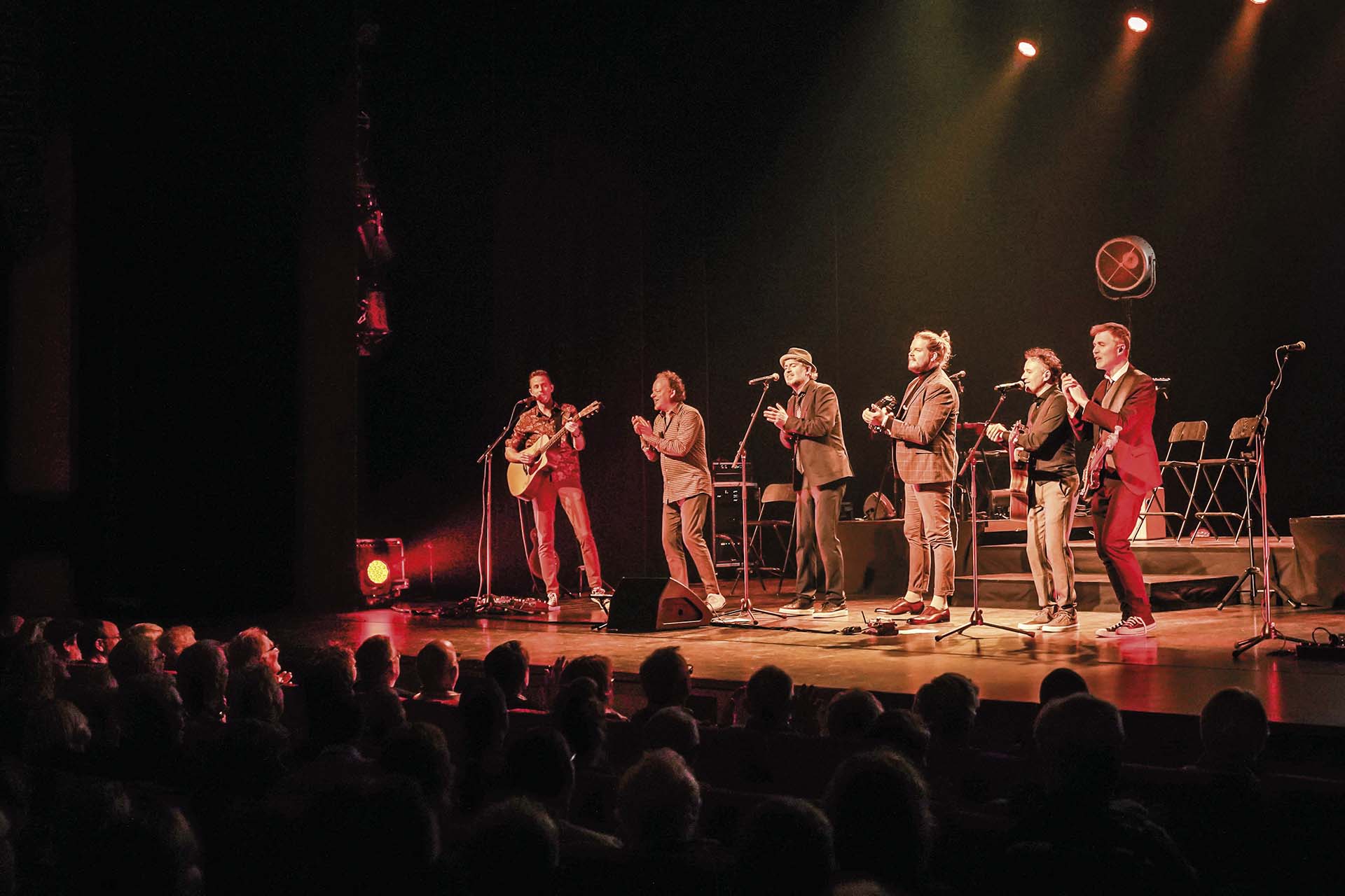De bandleden van Evergreen Top 1000 Live staan in een rij vooraan op het podium en zingen gezamenlijk voor een enthousiast publiek.