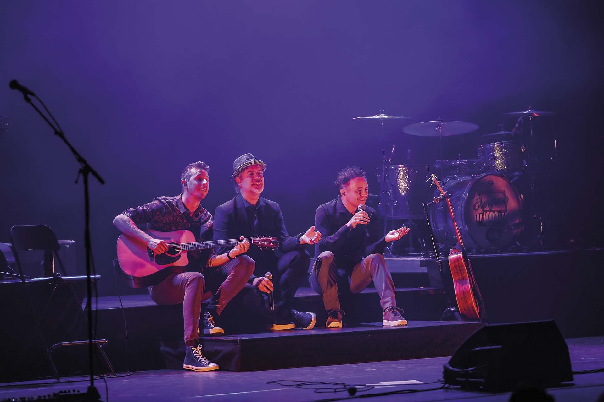 Drie bandleden van Evergreen Top 1000 Live zitten vooraan op het podium, één speelt gitaar, de anderen zingen en praten tegen het publiek.