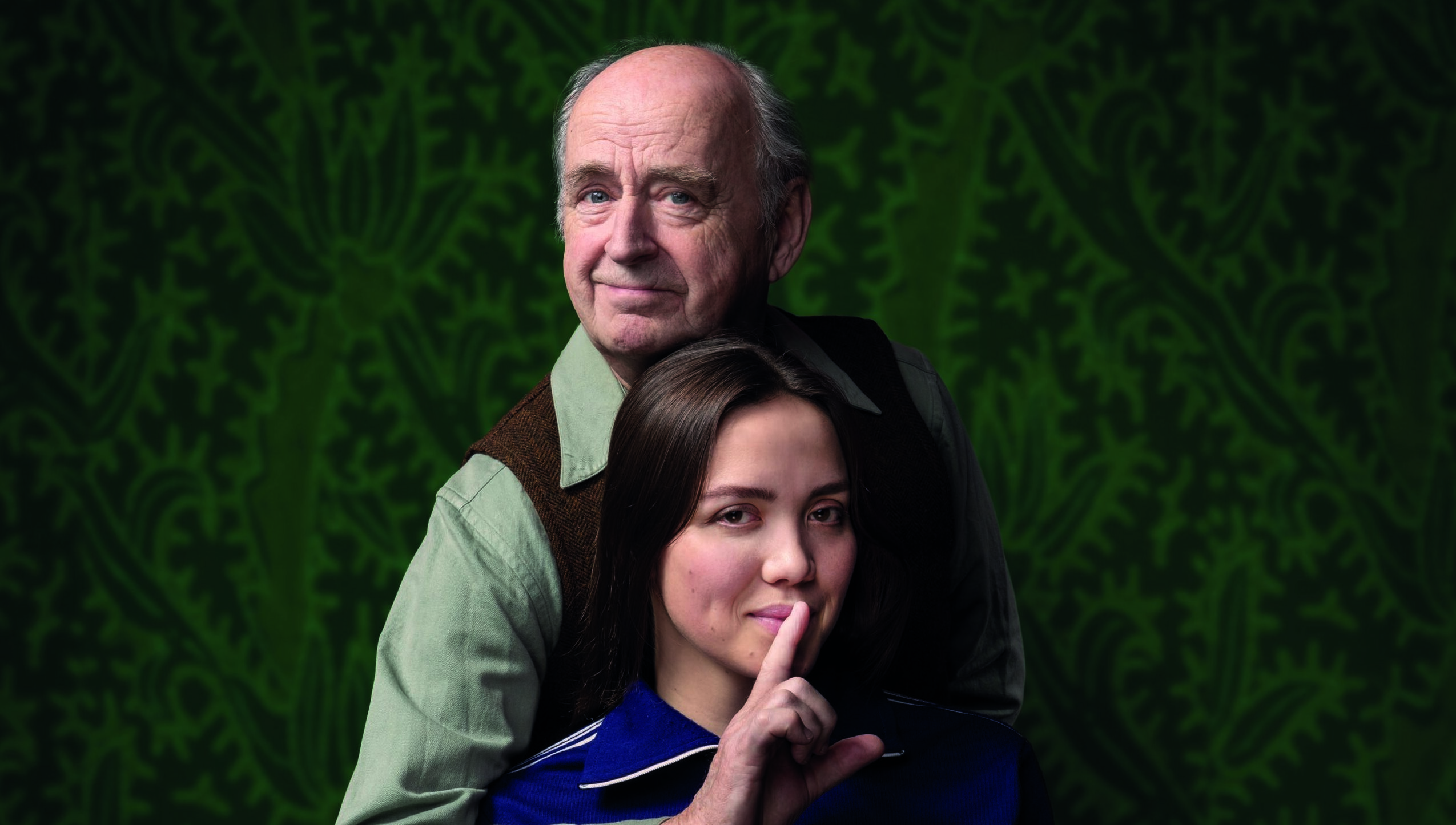 Oudere man maakt stilteggebaar bij jongere vrouw tegen groene achtergrond.