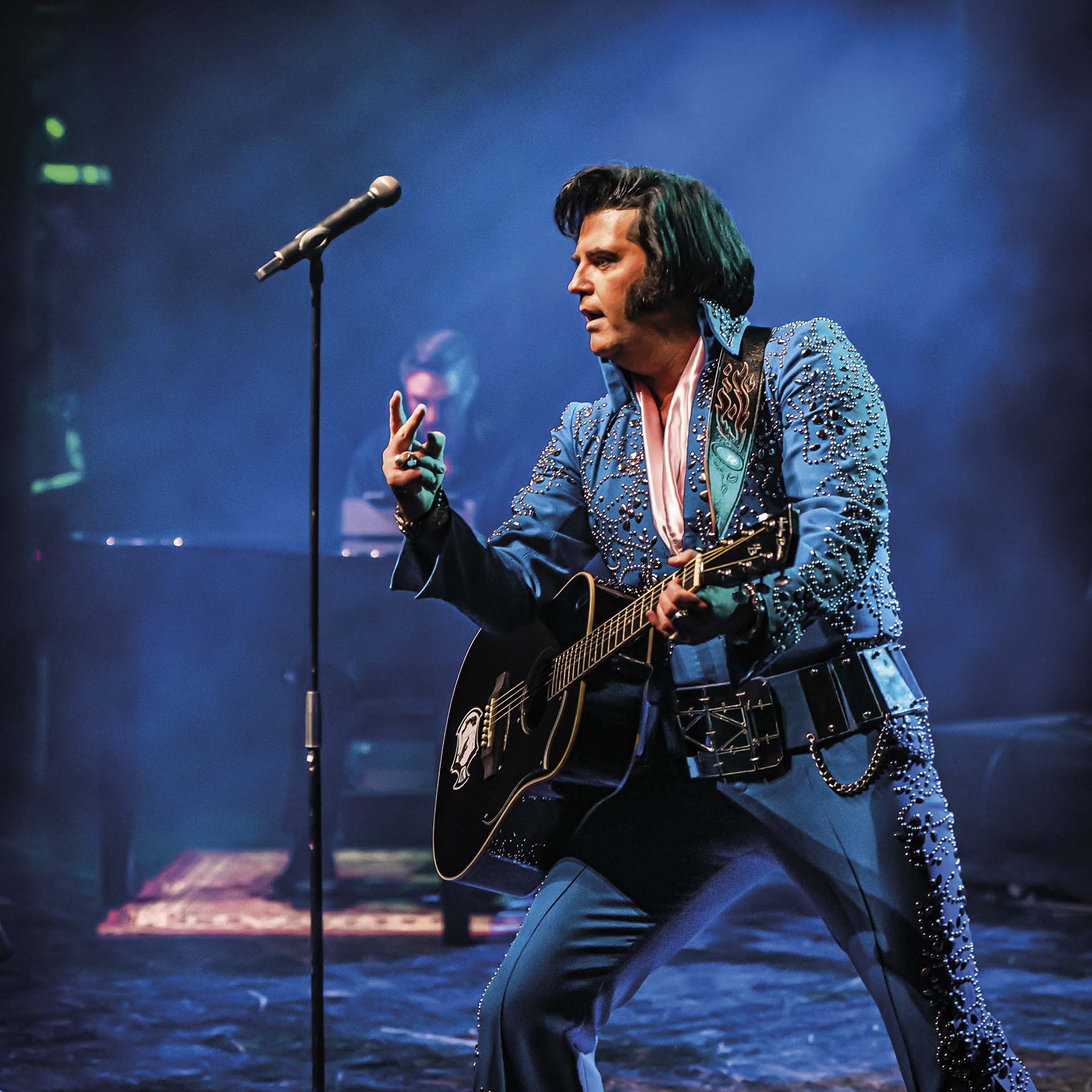 Zanger in Elvis-outfit speelt gitaar op podium met microfoon en blauwe spotlights.