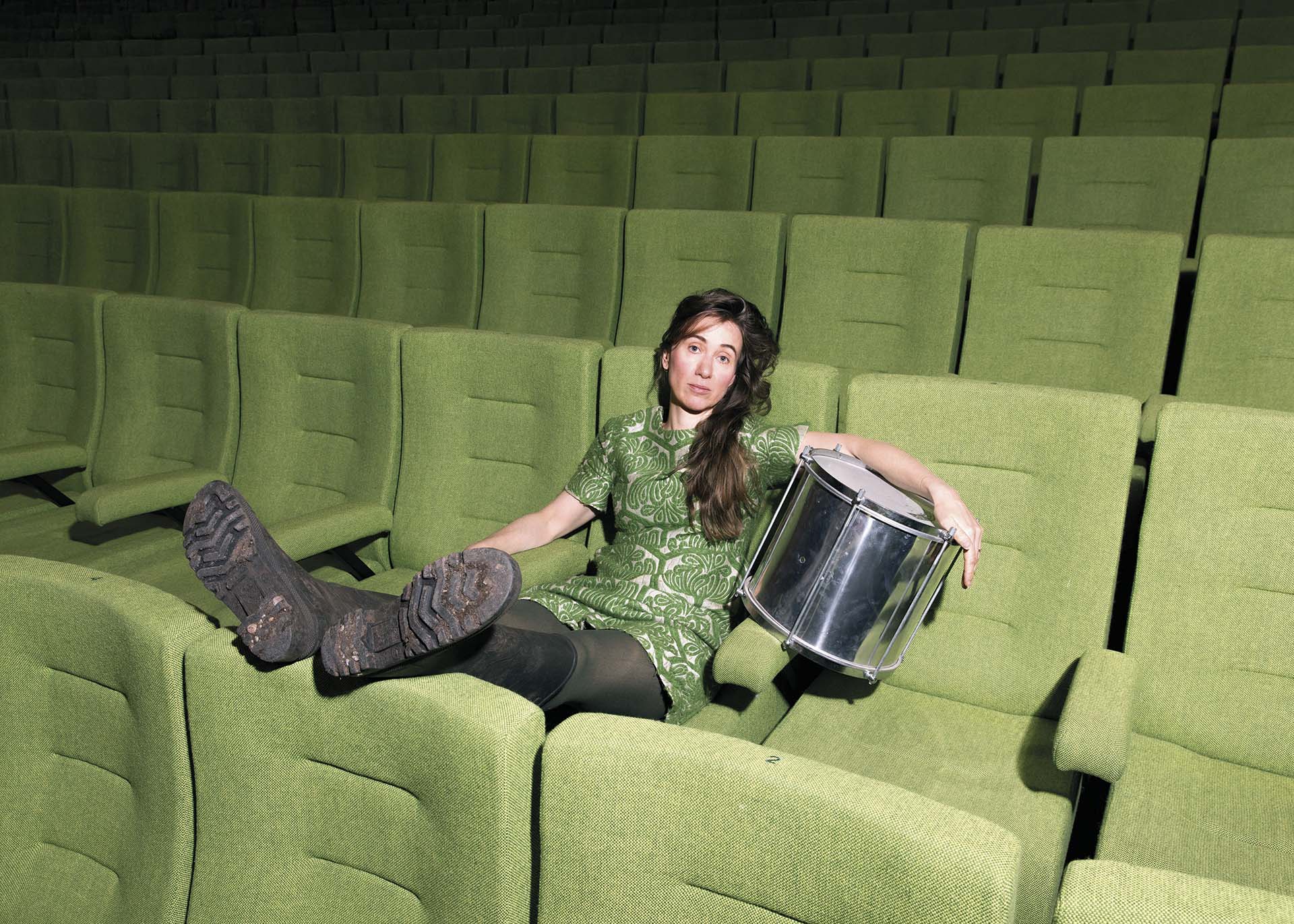Vrouw in groene jurk zit nonchalant met modderige laarzen op de stoelen van een lege theaterzaal, met een trommel op schoot.