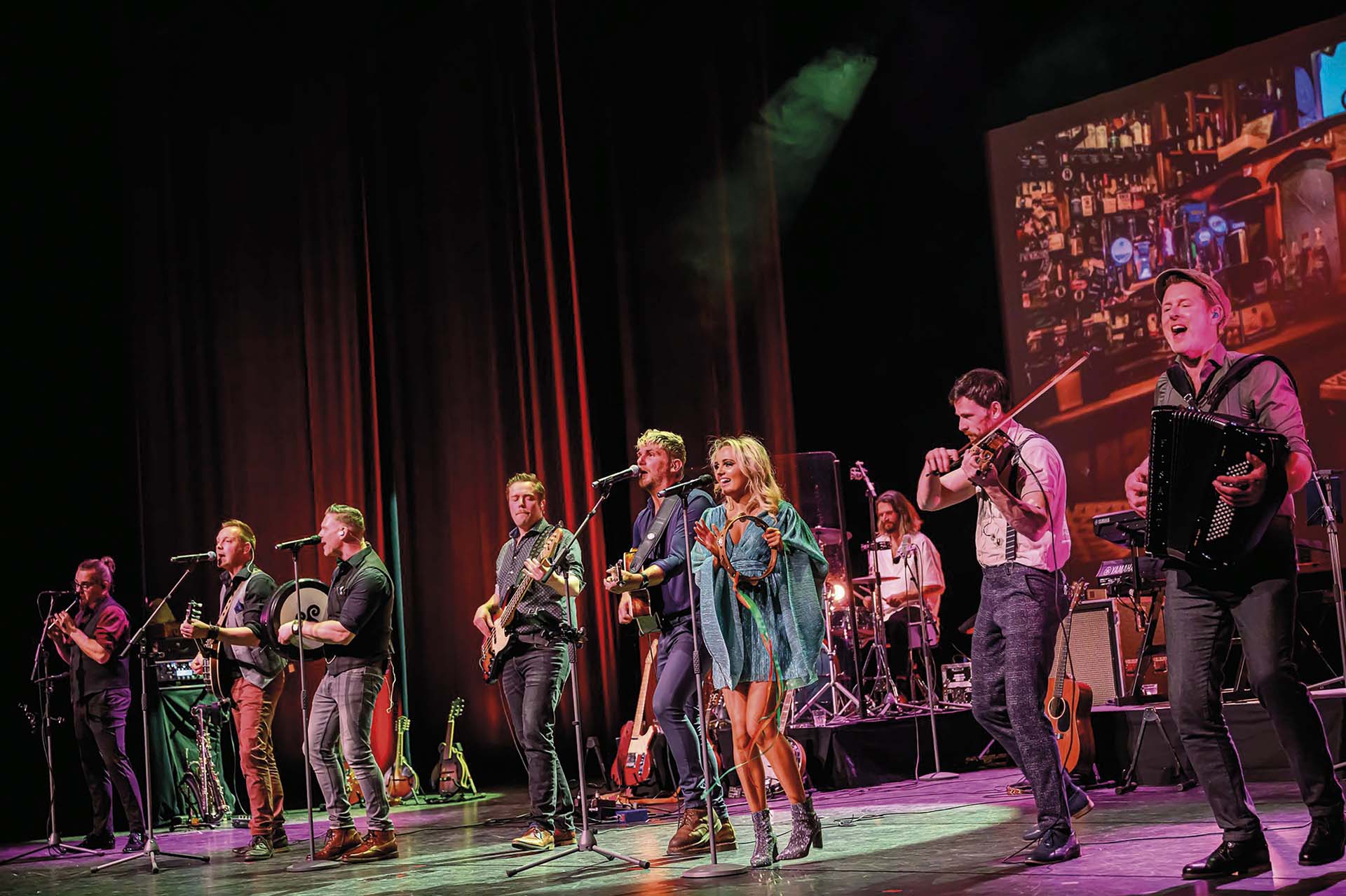 Grote muziekgroep met zang, viool, accordeon en gitaren treedt op op een theaterpodium, met kleurrijke belichting en een projectie van een bar op de achtergrond.