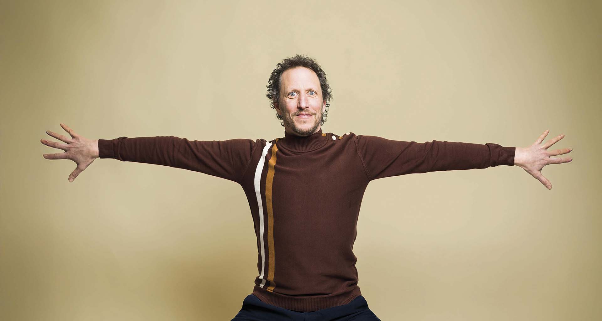 Micha Wertheim staat met uitgestrekte armen tegen een beige achtergrond, gekleed in een bruin shirt met verticale strepen.