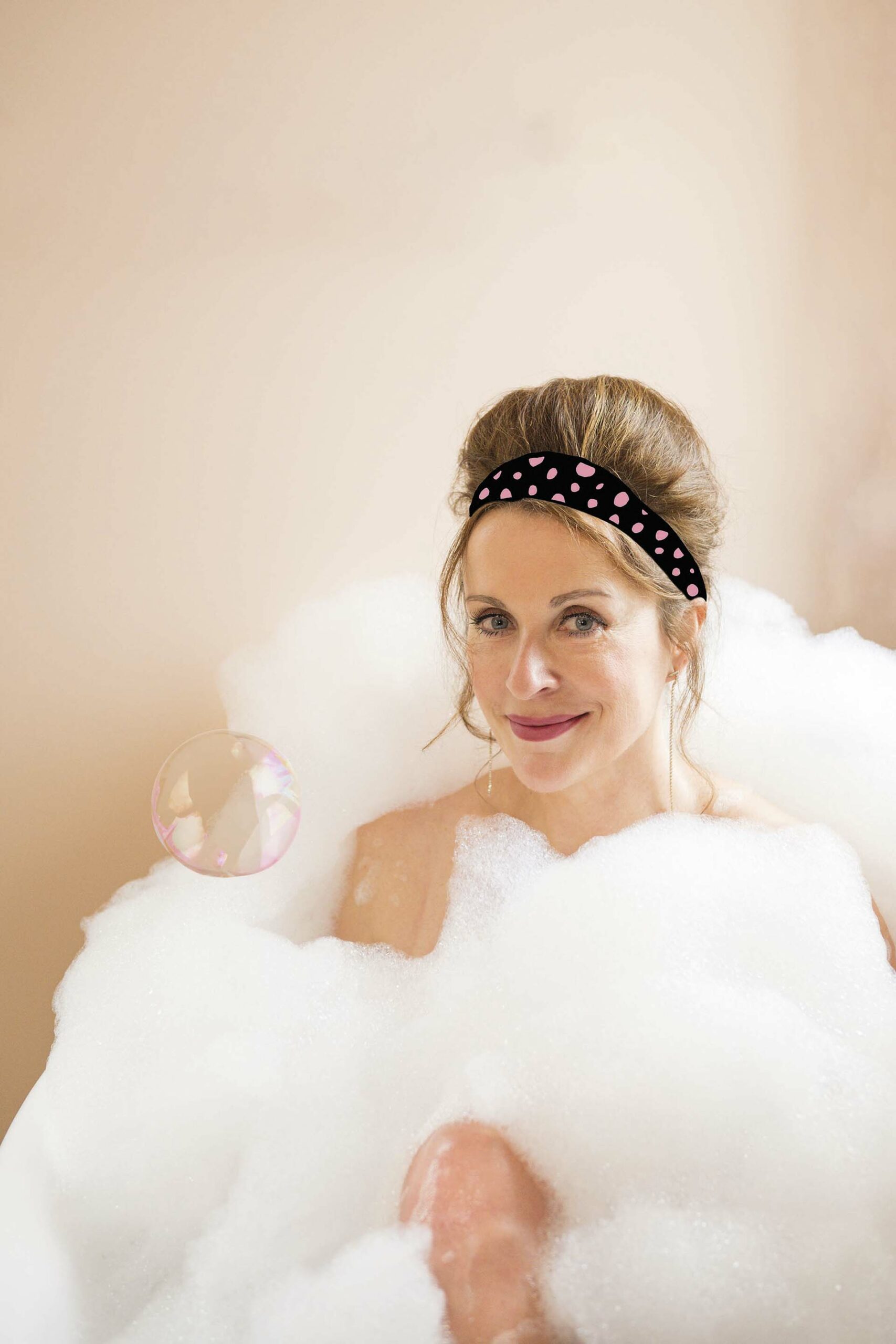 Annick Boer met opgestoken haar en roze haarband zit in een schuimbad en glimlacht naar de camera, met een zeepbel naast haar gezicht.