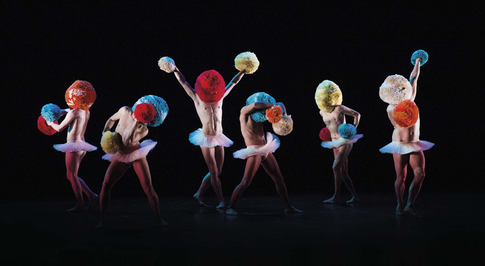 Zes dansers met kleurrijke pompons op hoofd en in handen voeren een choreografie uit in tutu’s.