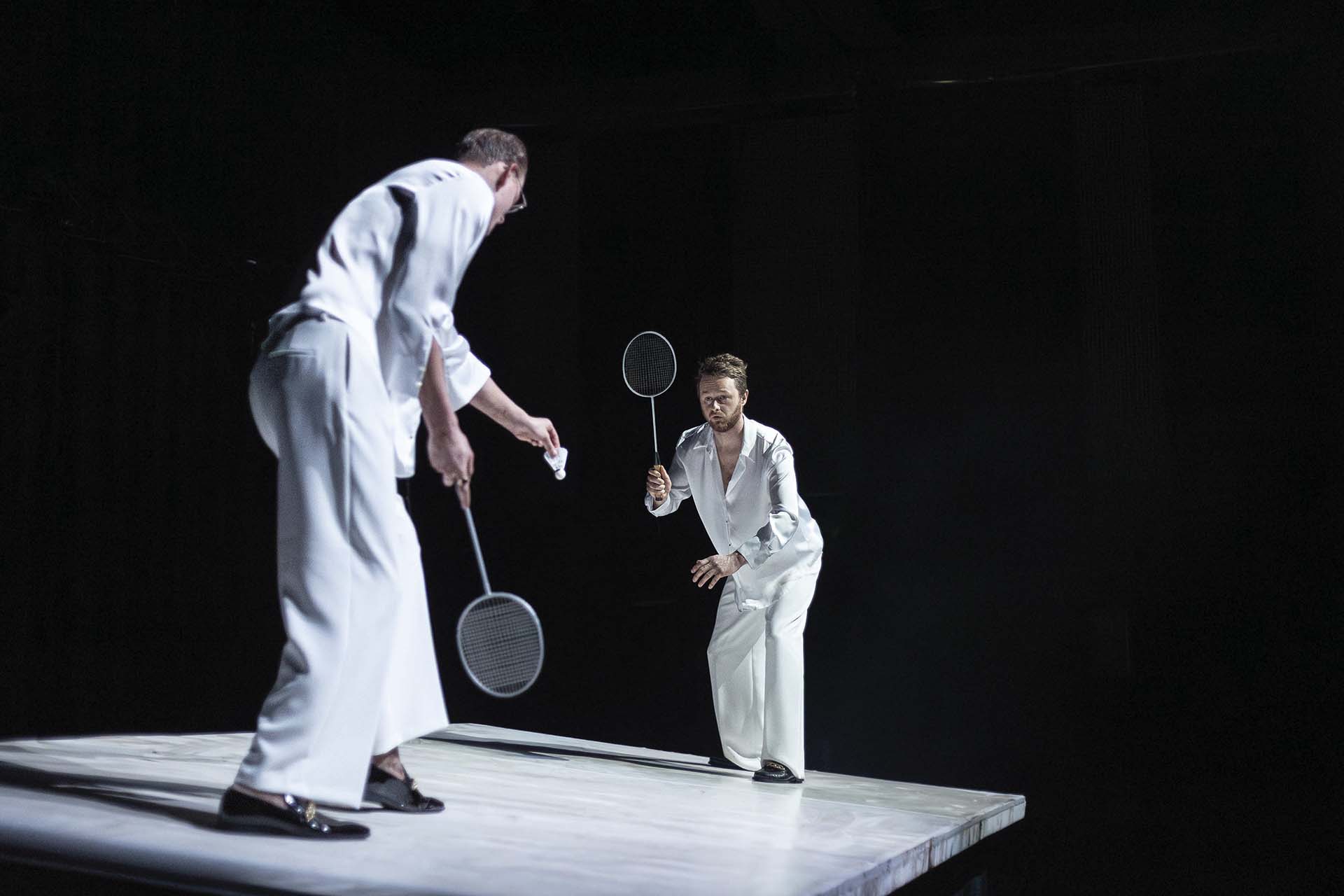 Twee mannen in witte kostuums spelen badminton op een verhoogd wit podium.