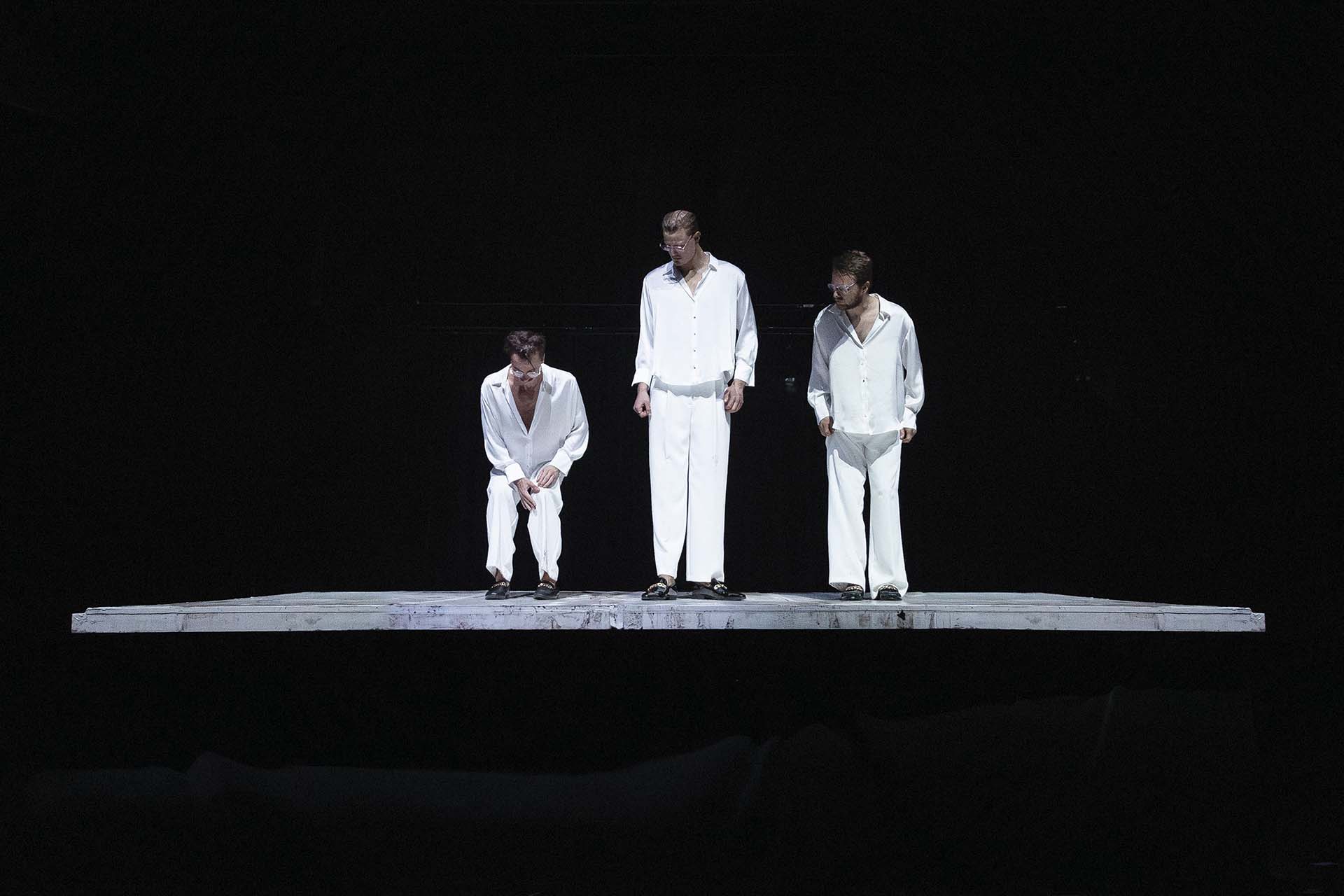 Drie mannen in witte kleding staan op een verlicht platform tegen een volledig zwarte achtergrond tijdens Rebirth part II. HEAVEN van Collectief Het Paradijs.