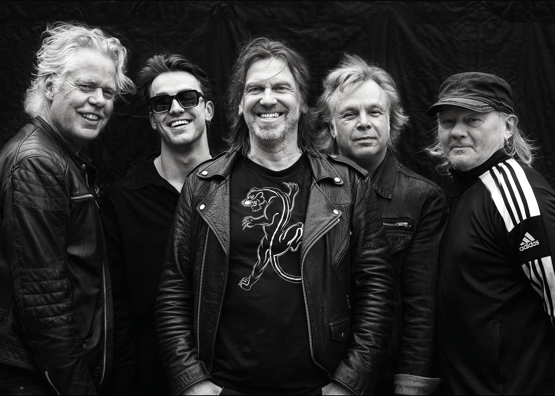 Groepsportret van de band Golden Earring Undercover in leren jacks en zwarte kleding, lachend in de camera voor een donkere achtergrond.