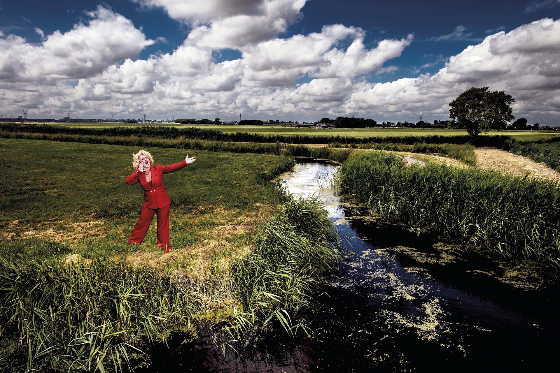 Karin Bloemen in rood pak roept in een weids Nederlands polderlandschap met sloot en dreigende wolkenlucht.
