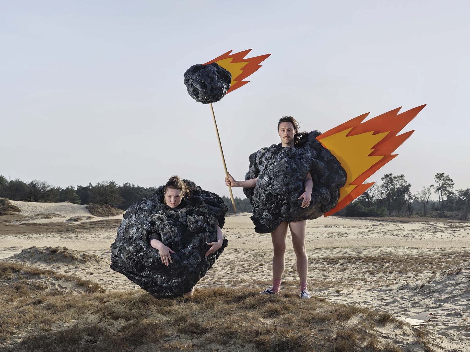 Twee performers in meteorietpakken met vlammen staan in een dor zandlandschap; één draagt vleugels van vuur, de ander kijkt berustend uit een bolvormig kostuum.