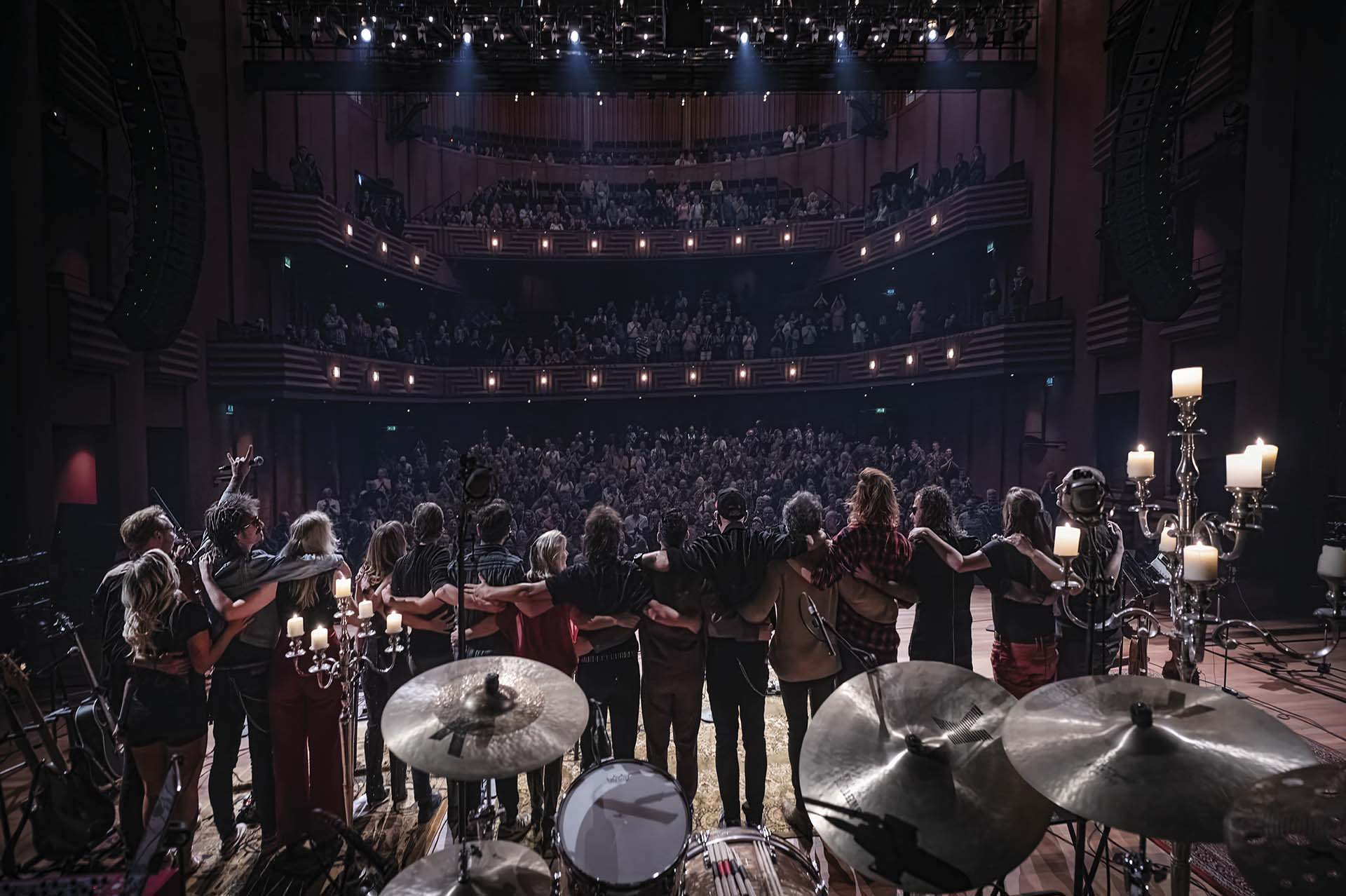 Muzikanten met de armen om elkaar heen buigen voor een staand publiek in een volle theaterzaal, gezien vanaf het podium tussen drums en kandelaars.
