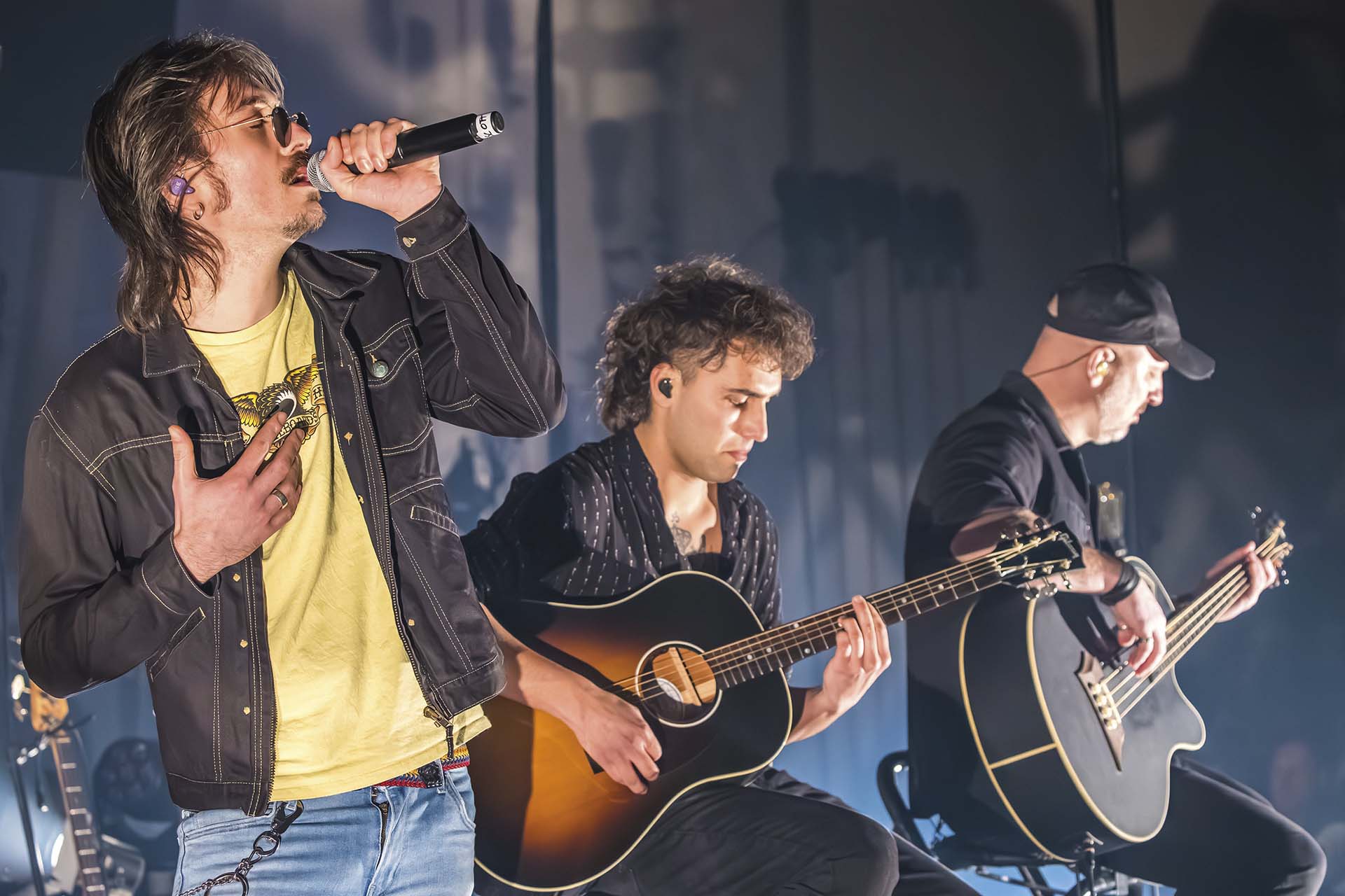 Zanger met microfoon en twee gitaristen tijdens een akoestisch optreden, als eerbetoon aan MTV Unplugged.