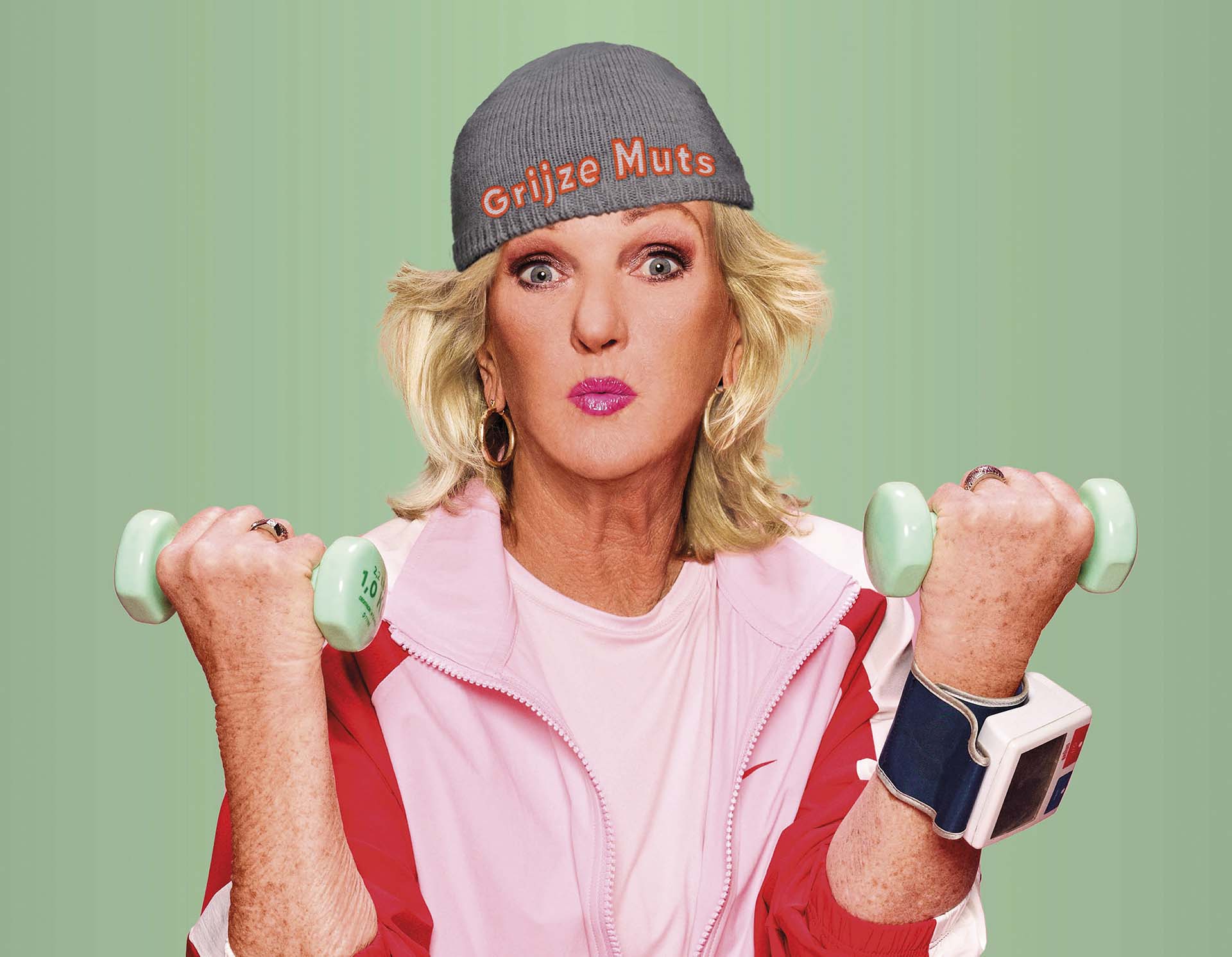 Vrouw met grijze muts en roze sportoutfit tilt gewichten tegen groene achtergrond.