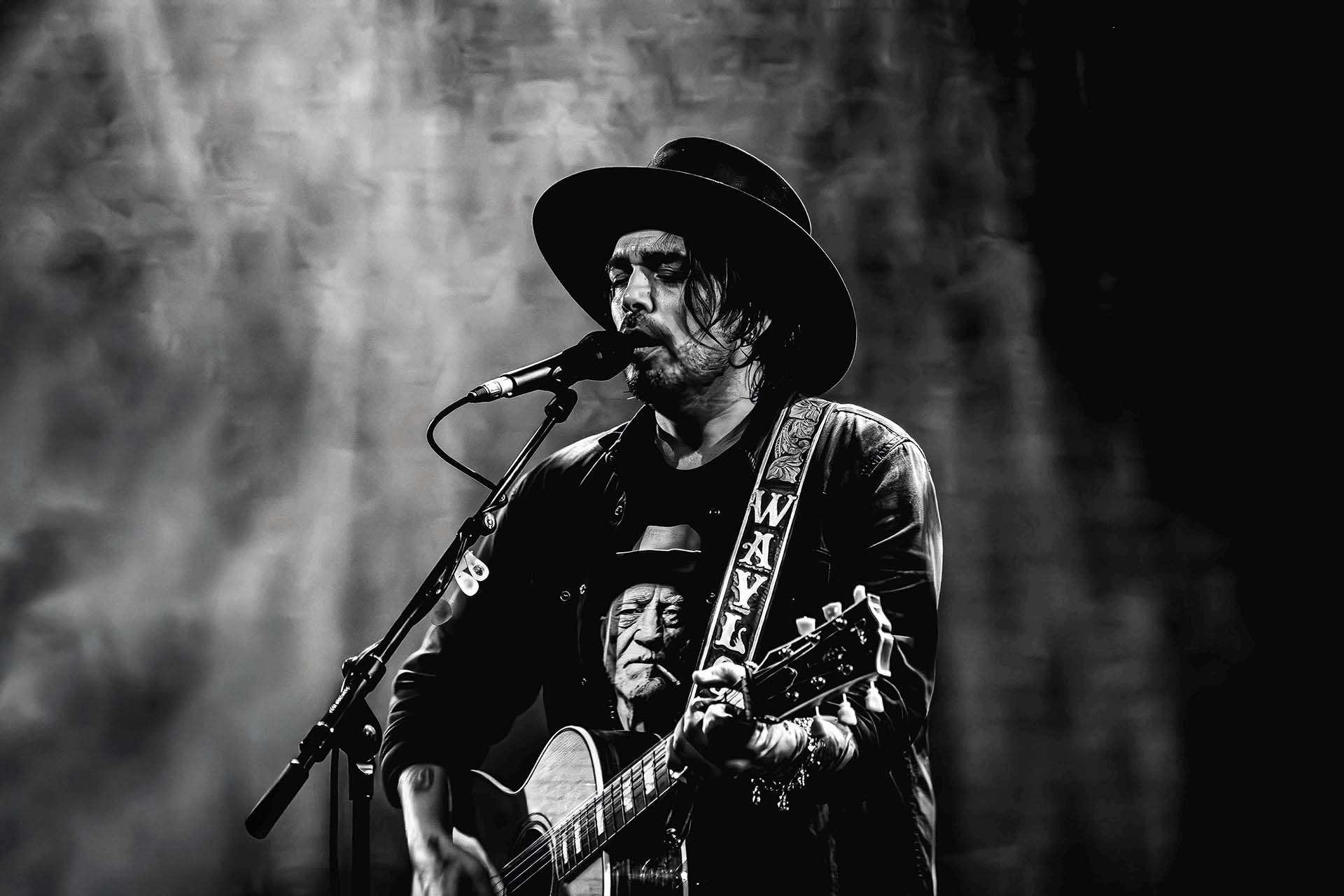 Waylon zingt in een microfoon en speelt gitaar tijdens een optreden, gekleed in een zwart leren jack en hoed, met het woord 'WAYLON' op de gitaarband, in zwart-wit vastgelegd.