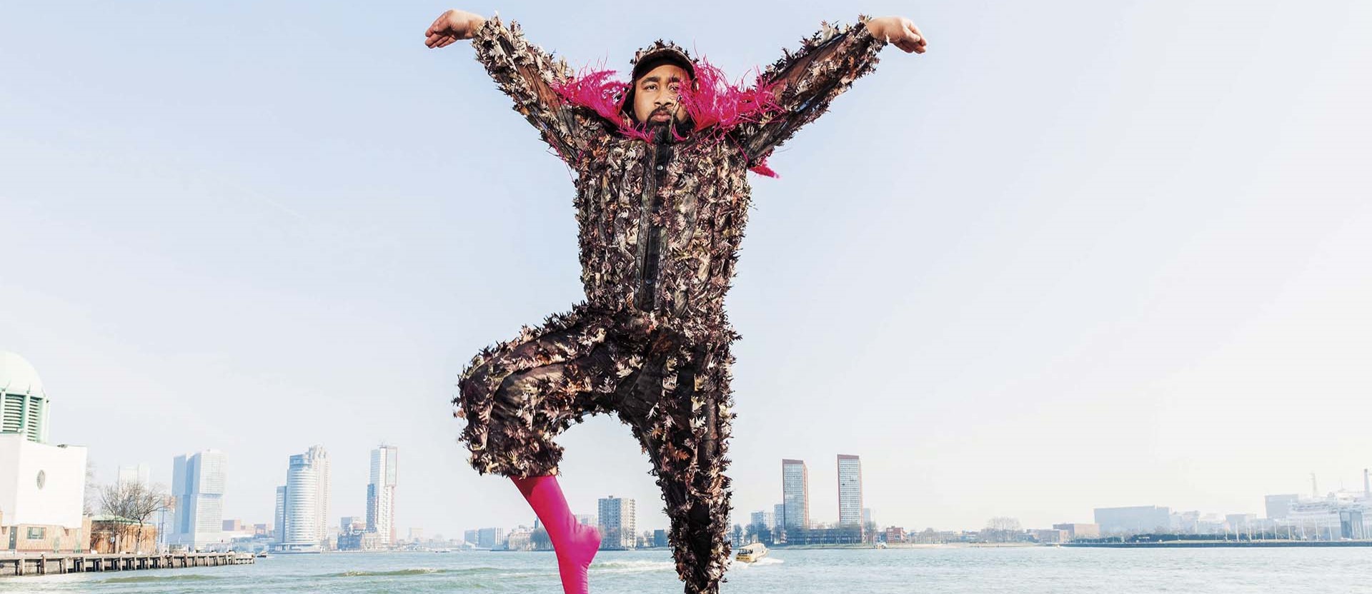 Een performer in een pak met bladeren en roze details springt hoog in de lucht voor een skyline aan het water.