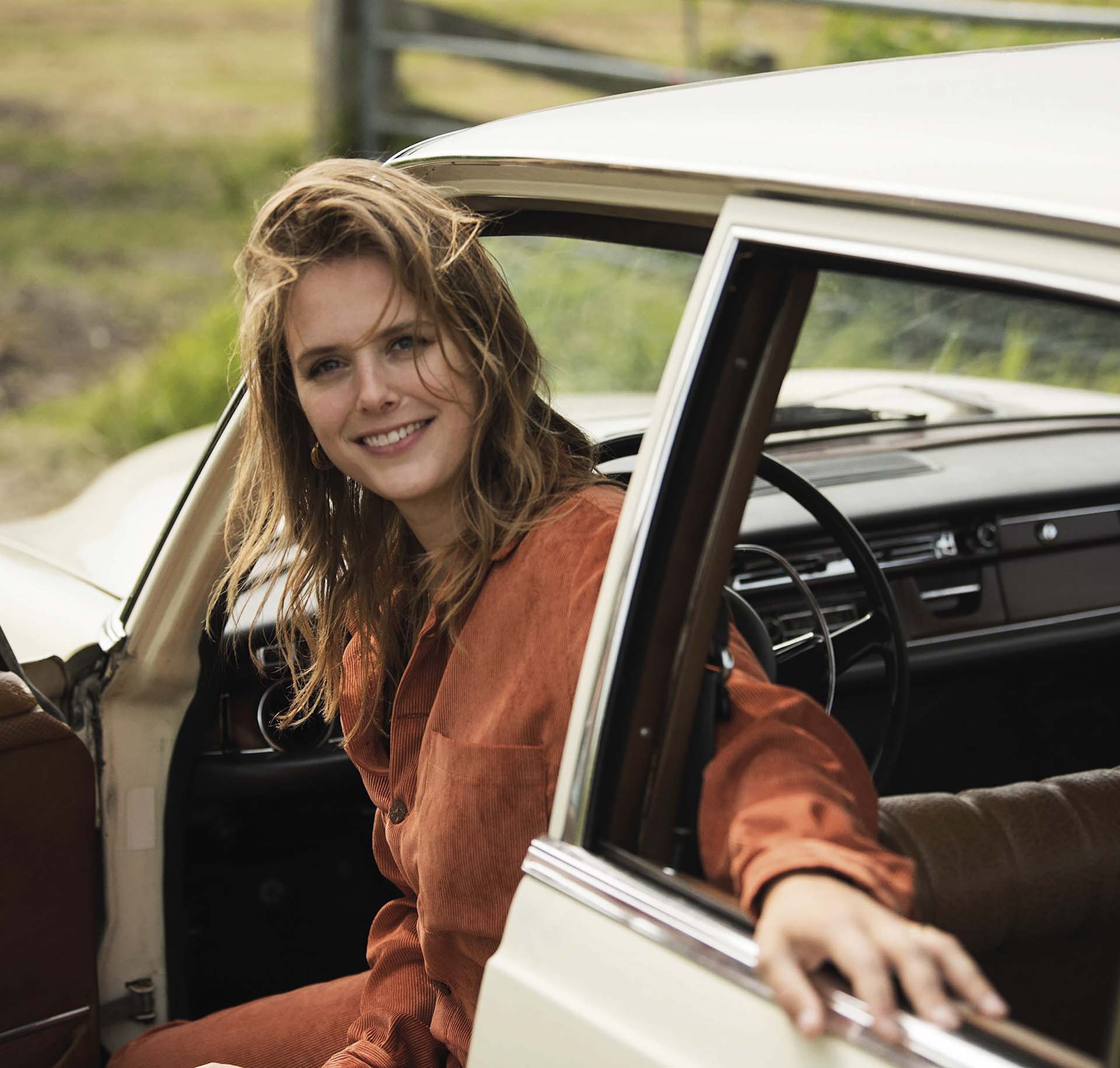 Maaike Ouboter zit in een openstaande autodeur van een vintage auto, glimlachend naar de camera in een natuurlijke, zomerse omgeving.