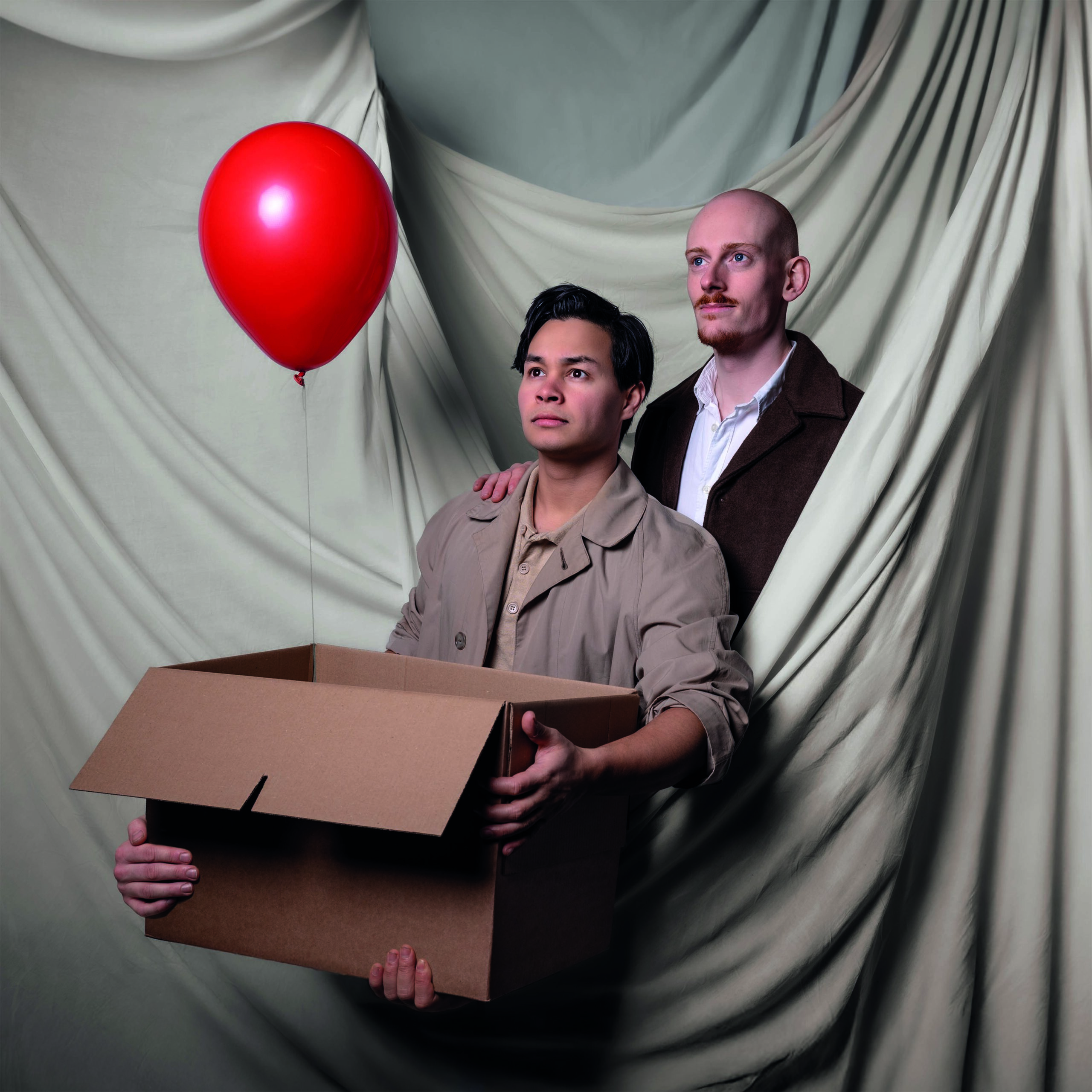 Niek en Guy staan voor een gordijnachtergrond, één met een rode ballon en de ander met een grote kartonnen doos, kijken serieus in de camera.