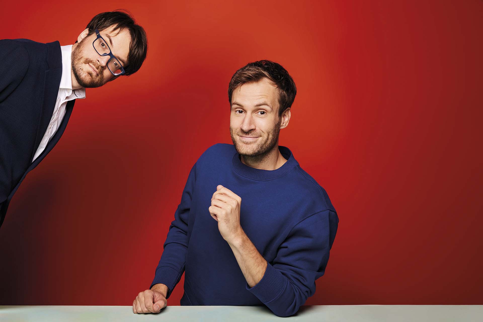 Roel en Jos poseren voor rode achtergrond, één met bril en de ander glimlachend.