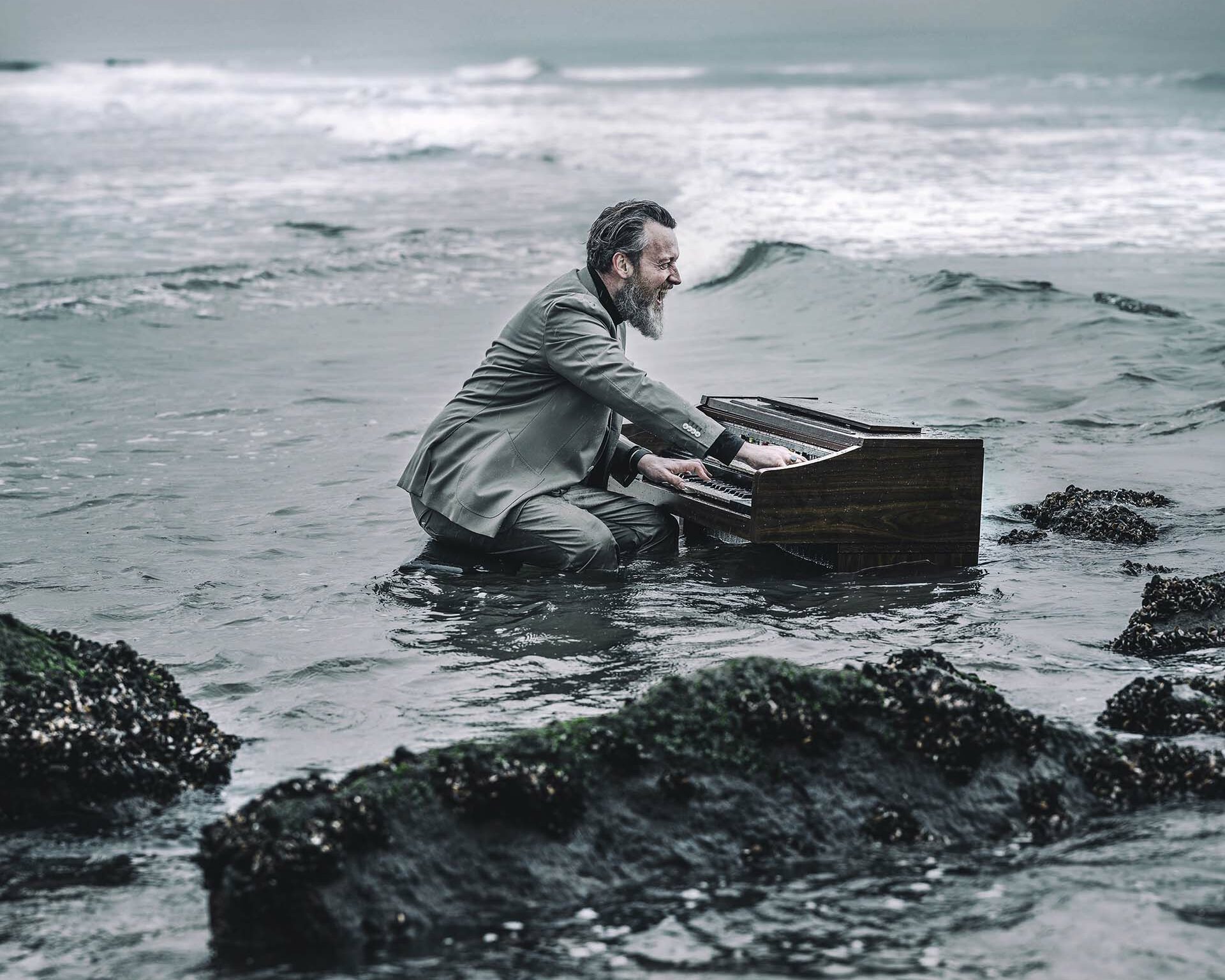 Een man in pak knielt in de branding en speelt piano in zee, terwijl dreigende wolken boven de woeste Noordzee hangen.