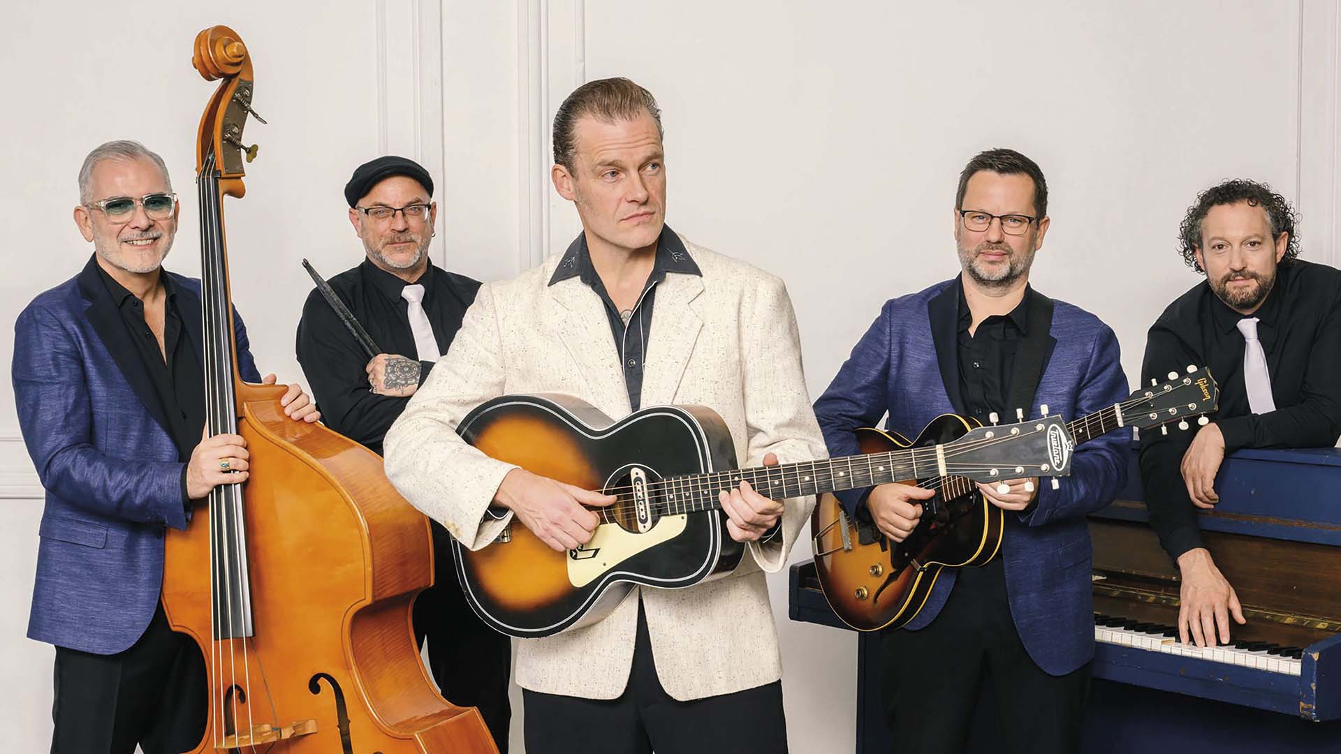 The Wanderers poseren met contrabas, gitaar en piano, met de zanger centraal en instrumenten in de hand.