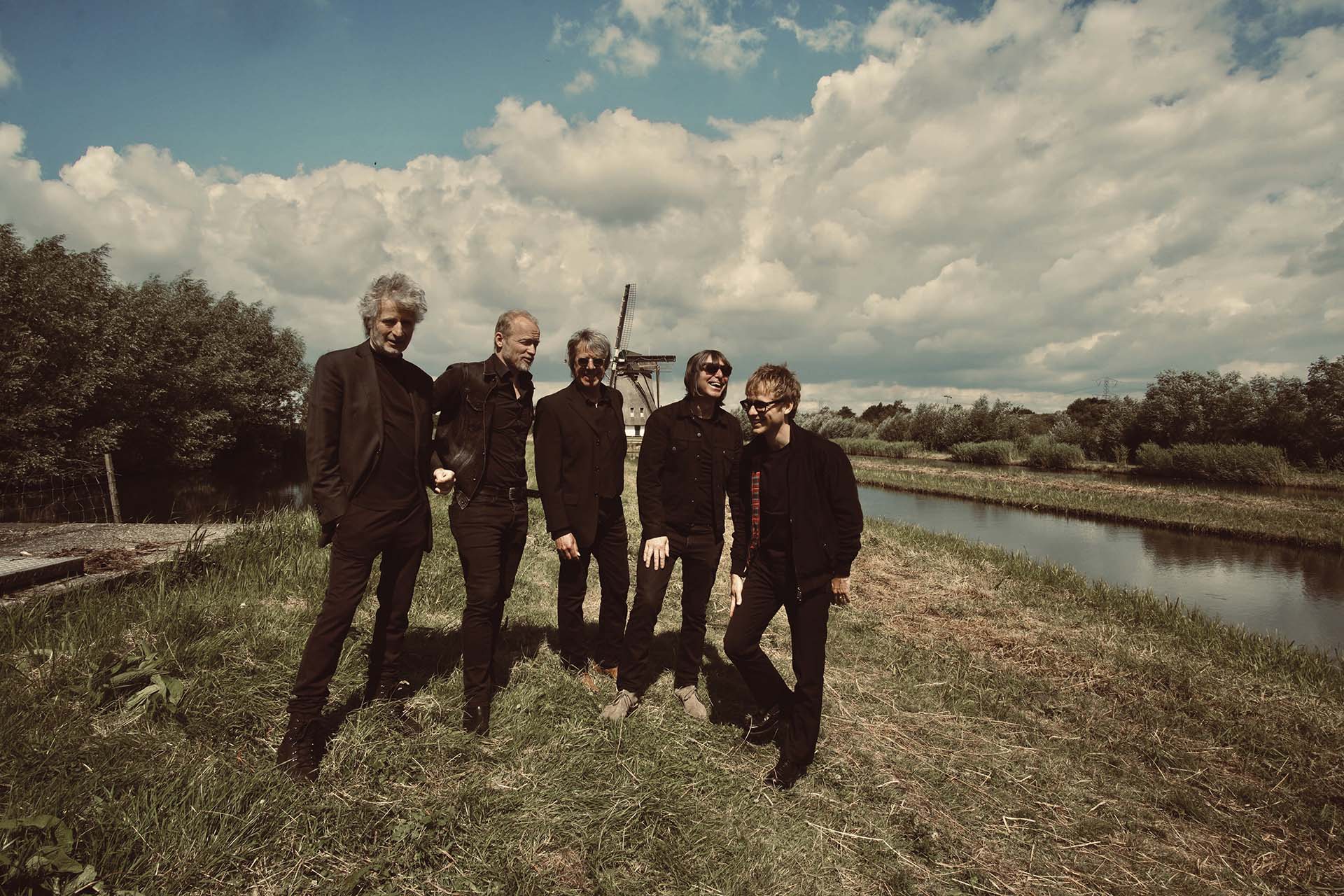 De bandleden van Timebox staan lachend in het gras langs een watergang, met op de achtergrond een molen en een wolkenlucht.