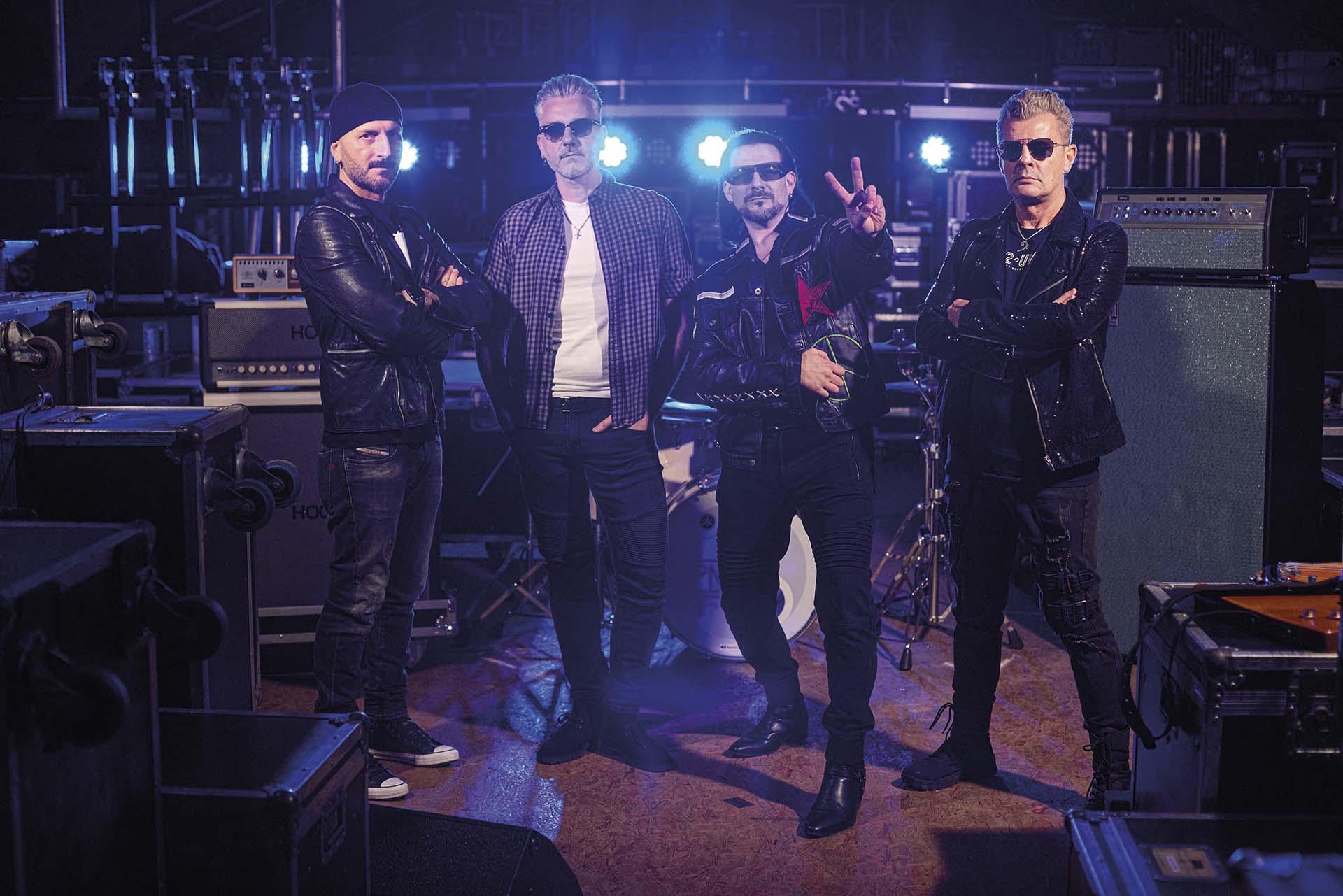 Bandleden van U2 THE MUSIC poseren in een donkere studio met versterkers op de achtergrond, allemaal in leren outfits en zonnebrillen, klaar voor hun tribute-optreden.