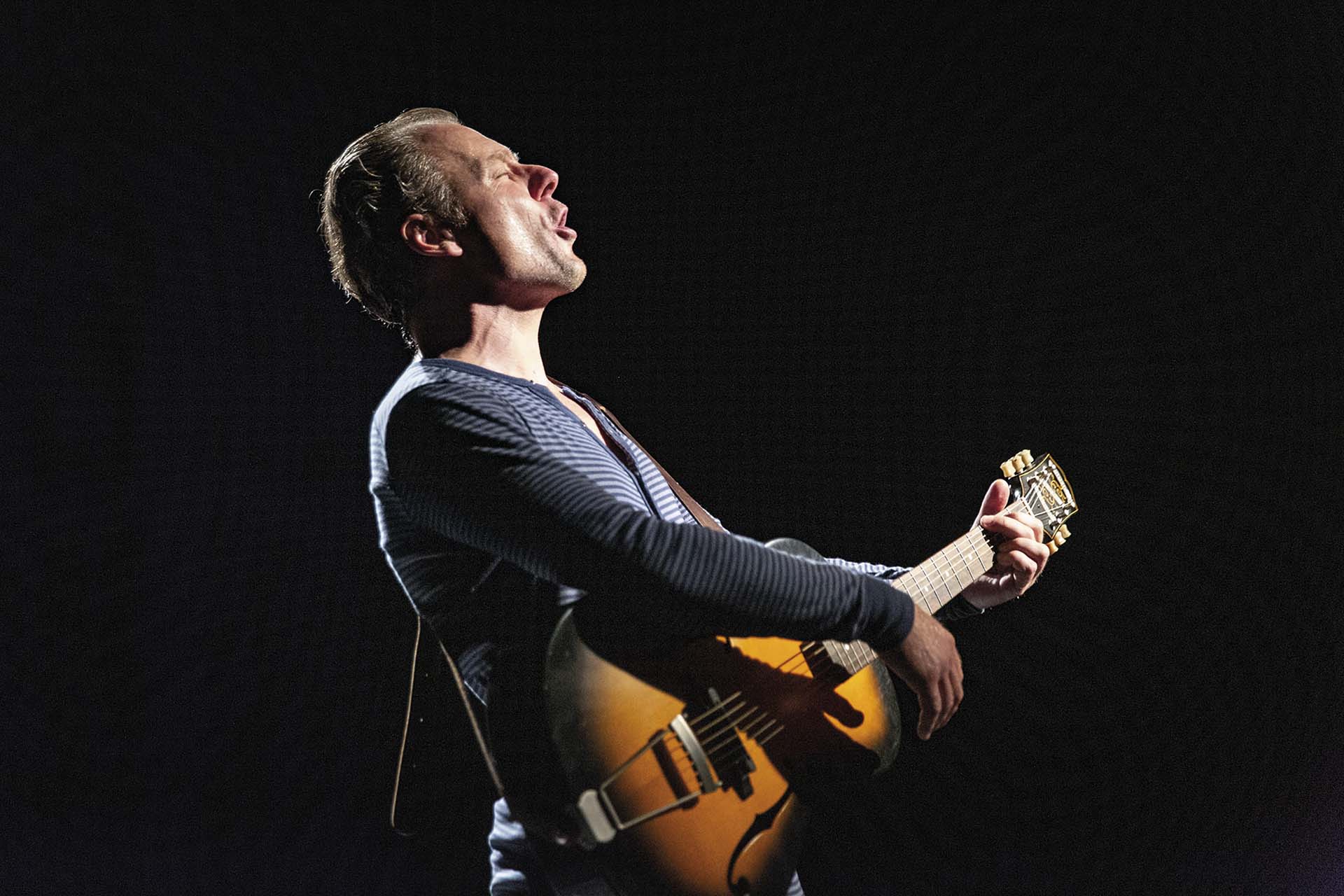 Viktor Griffioen met gitaar zingt gepassioneerd, verlicht op een donkere theaterachtergrond.