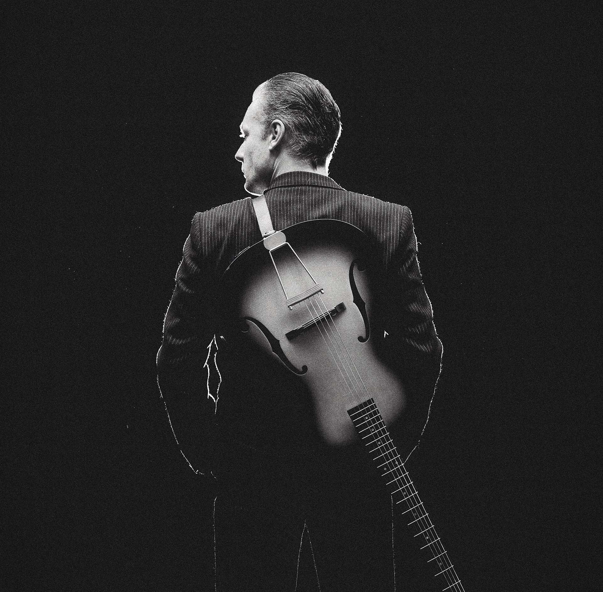 Viktor Griffioen staat in pak met zijn rug naar de camera, een gitaar op zijn rug, tegen een donkere achtergrond.
