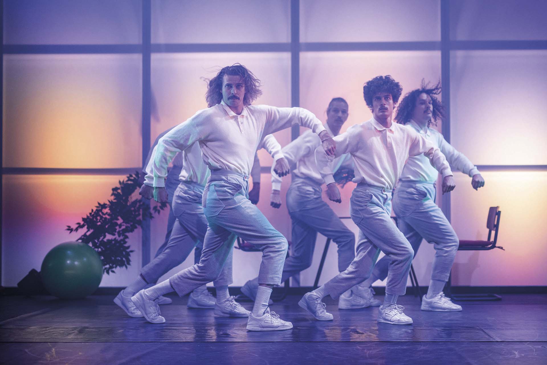Vijf dansers in witte outfits bewegen synchroon over podium met gekleurde lichtvlakken op de achtergrond, energieke choreografie in groepsformatie
