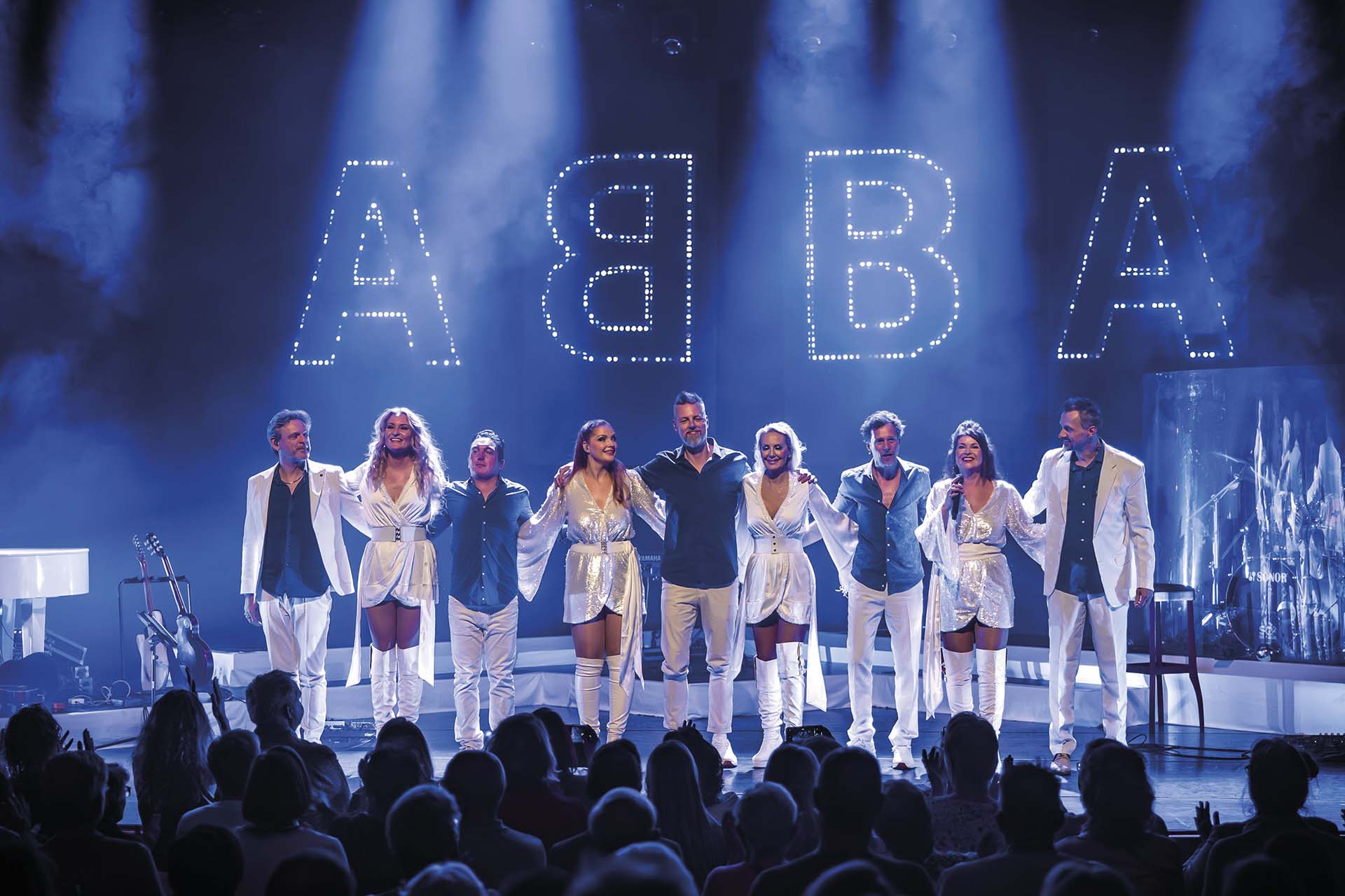 Groep performers in witte outfits buigt samen op het podium voor een applaudisserend publiek, met verlichte letters ABBA op de achtergrond
