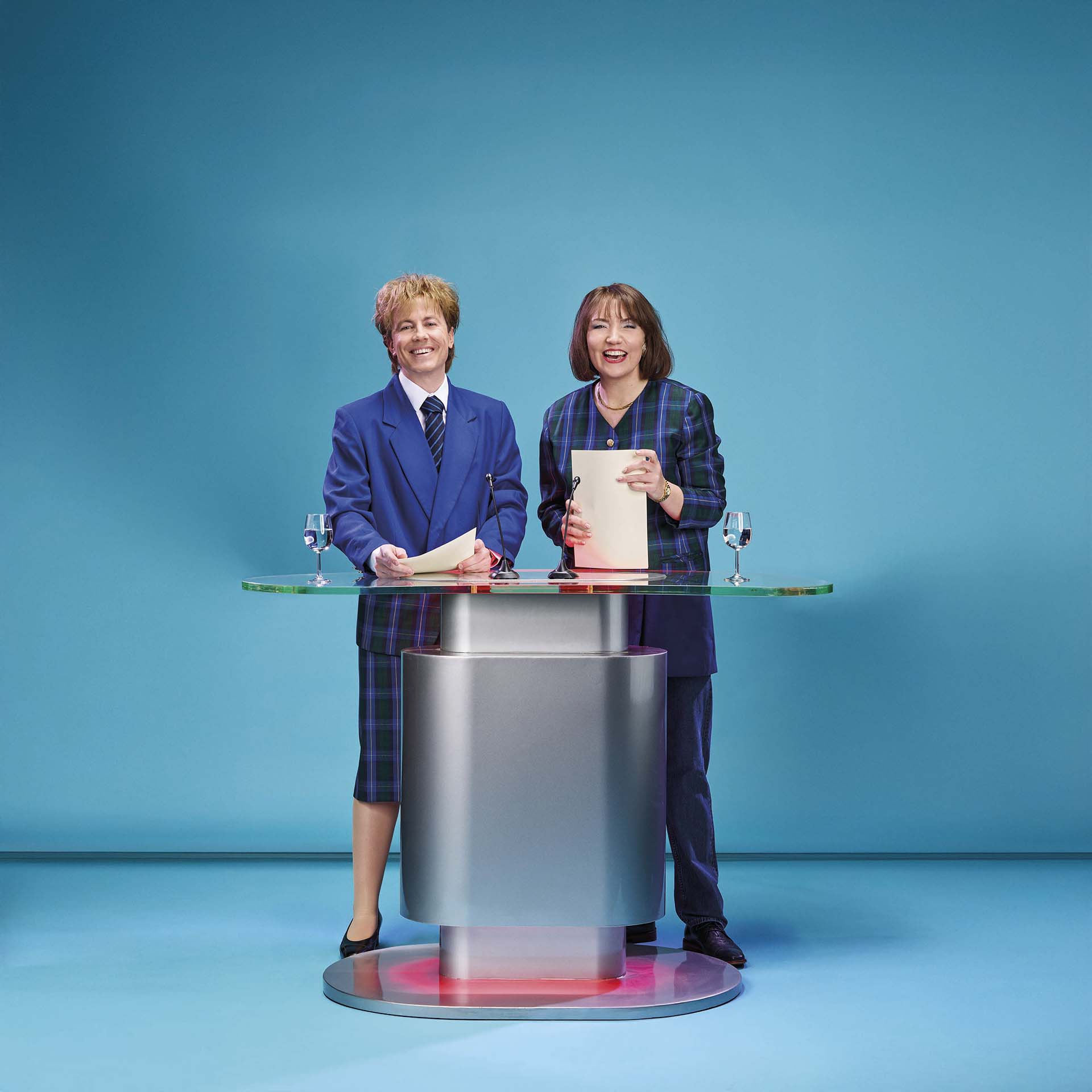Twee presentatoren staan achter een modern spreekgestoelte met papieren en glazen water tegen een blauwe achtergrond