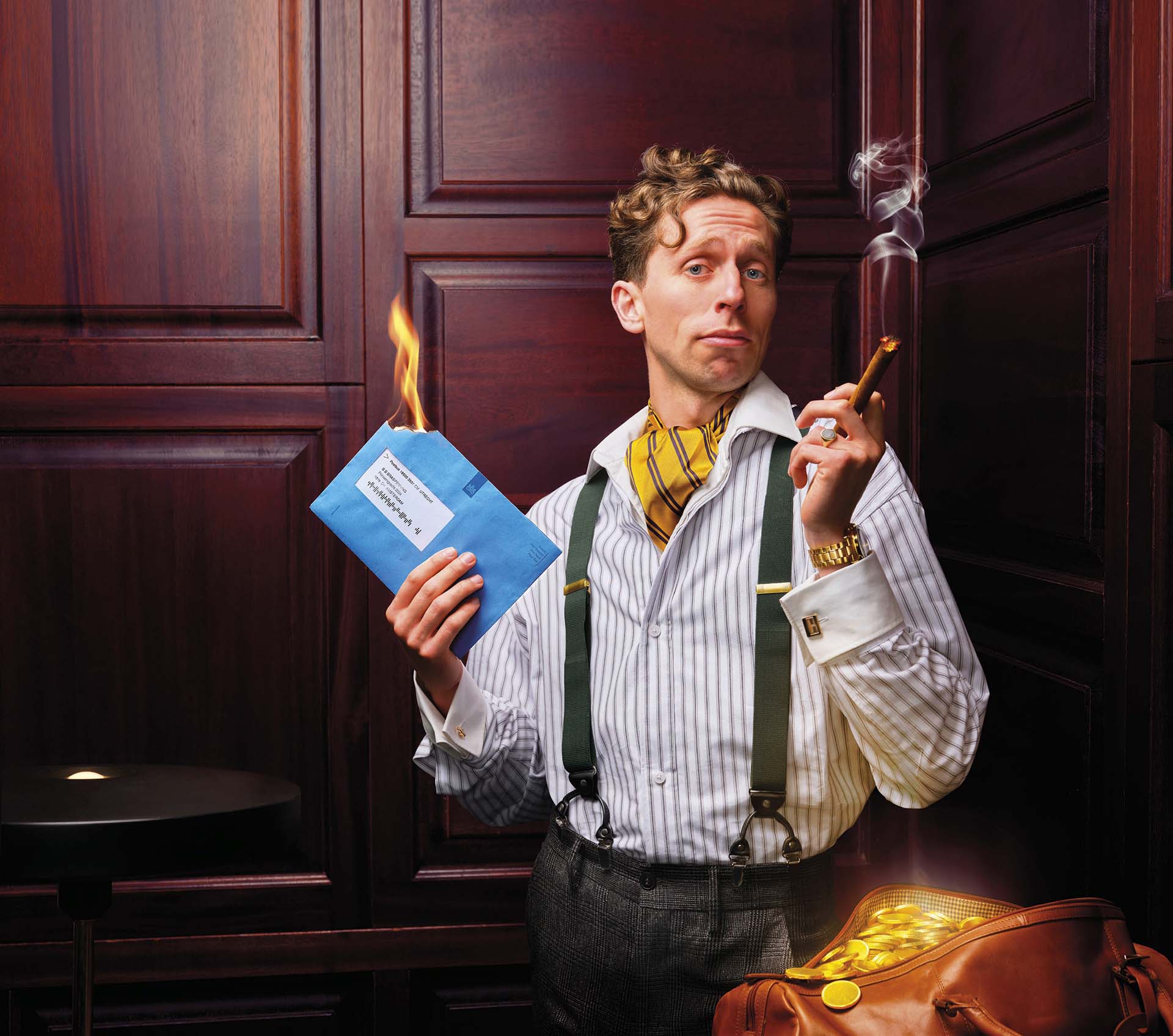 Boban Braspenning met bretels houdt brandende envelop en sigaar vast, naast open koffer met gouden munten tegen houten wand