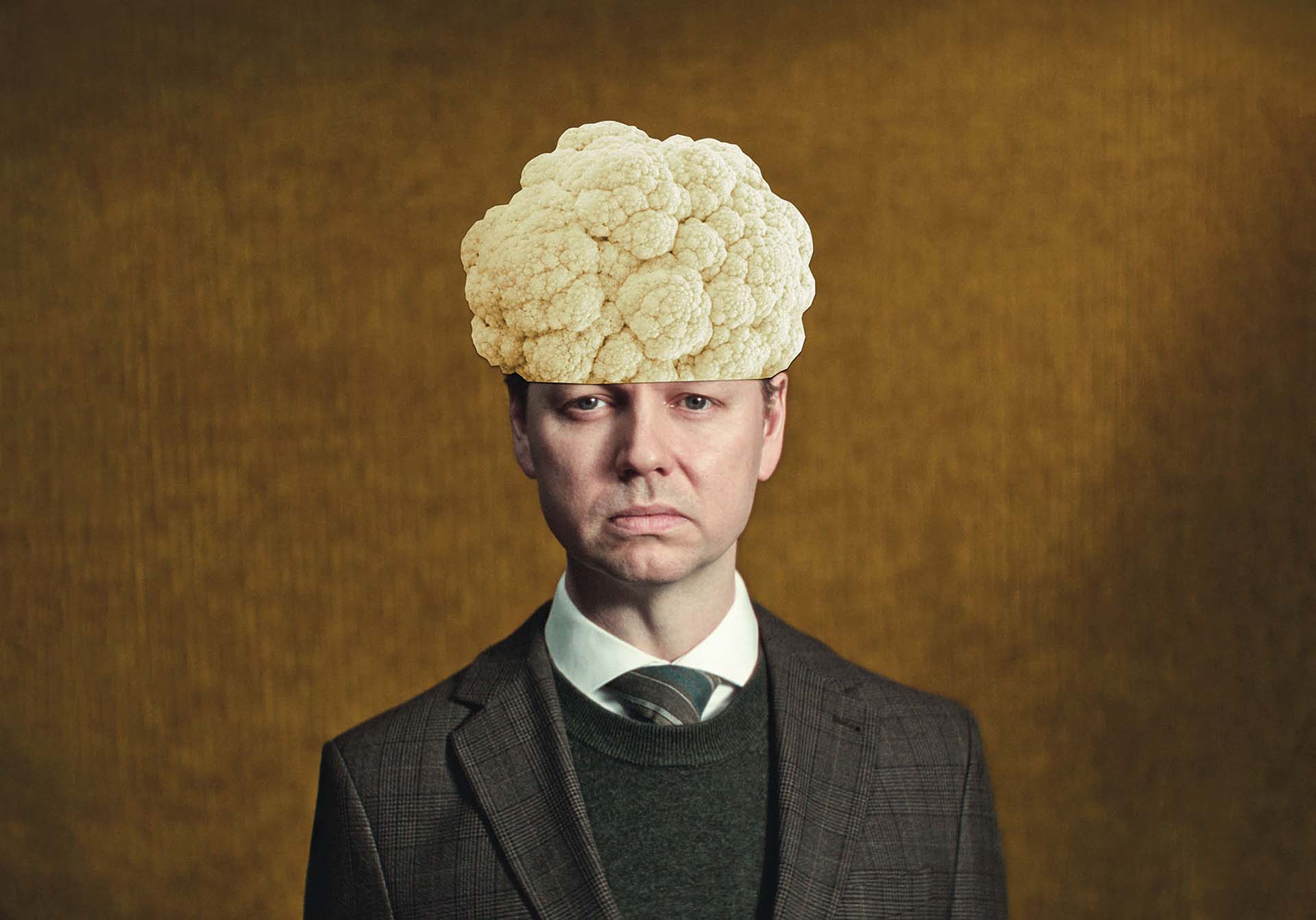Man in pak kijkt recht vooruit met een bloemkool op zijn hoofd tegen een warme, bruine achtergrond