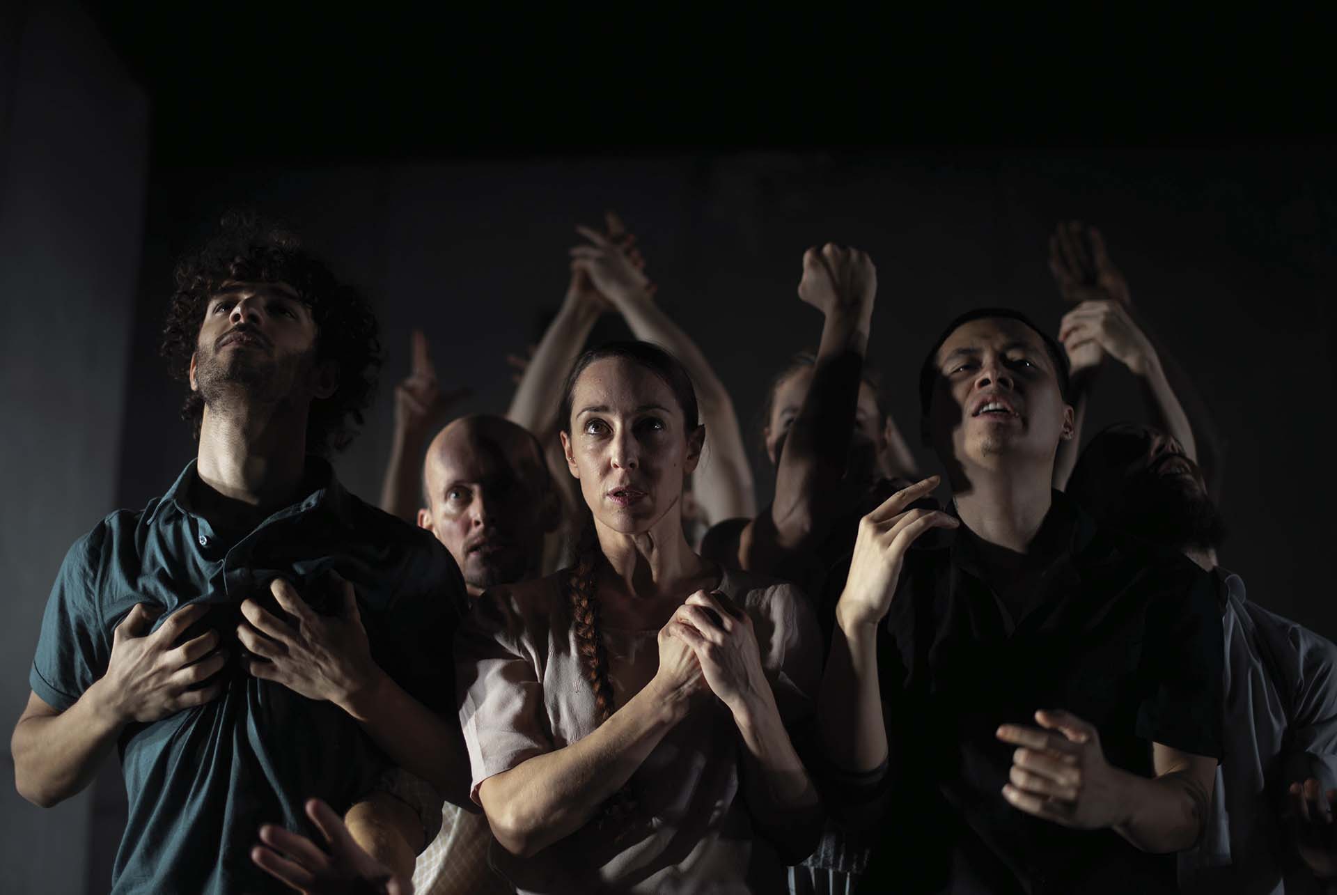Groep performers in donker licht op toneel, met expressieve blikken en opgeheven armen, alsof ze reageren op iets buiten beeld, in een intense en theatrale scène