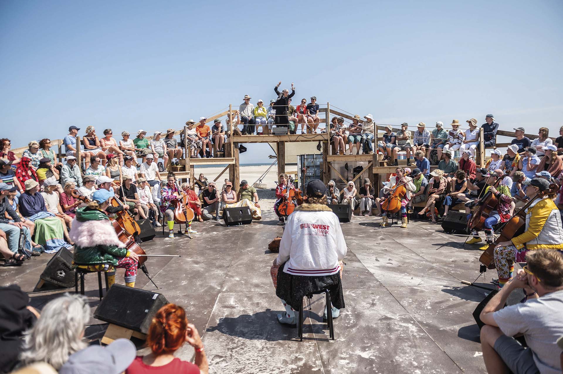 Publiek zit rondom een openluchtpodium op het strand terwijl een groep cellisten in een cirkel speelt, met een dirigent centraal op een verhoogd platform.