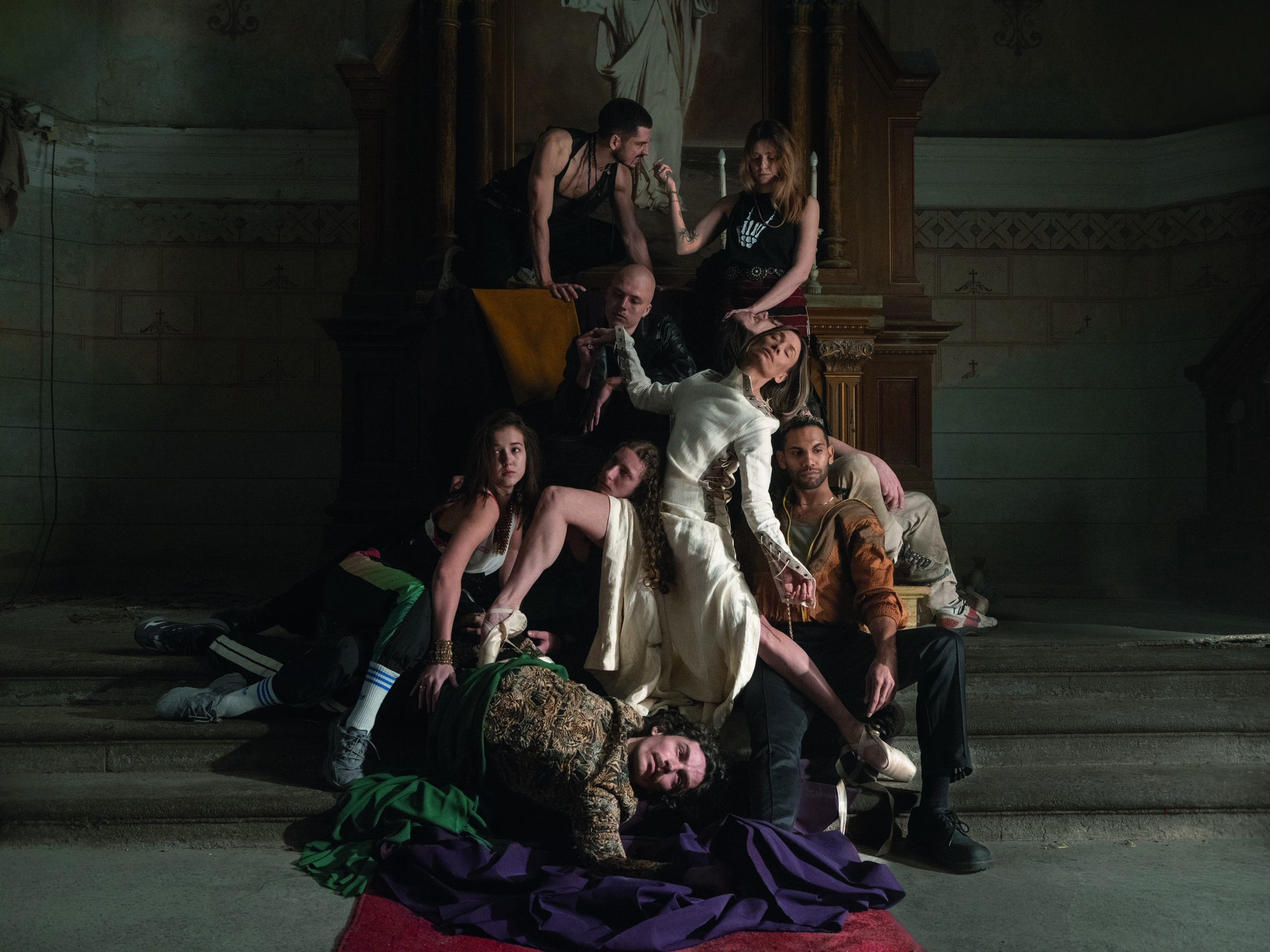 Groep dansers poseert in theatrale compositie op trappen in een klassiek interieur, met centrale vrouw in witte outfit en anderen daaromheen in expressieve houdingen