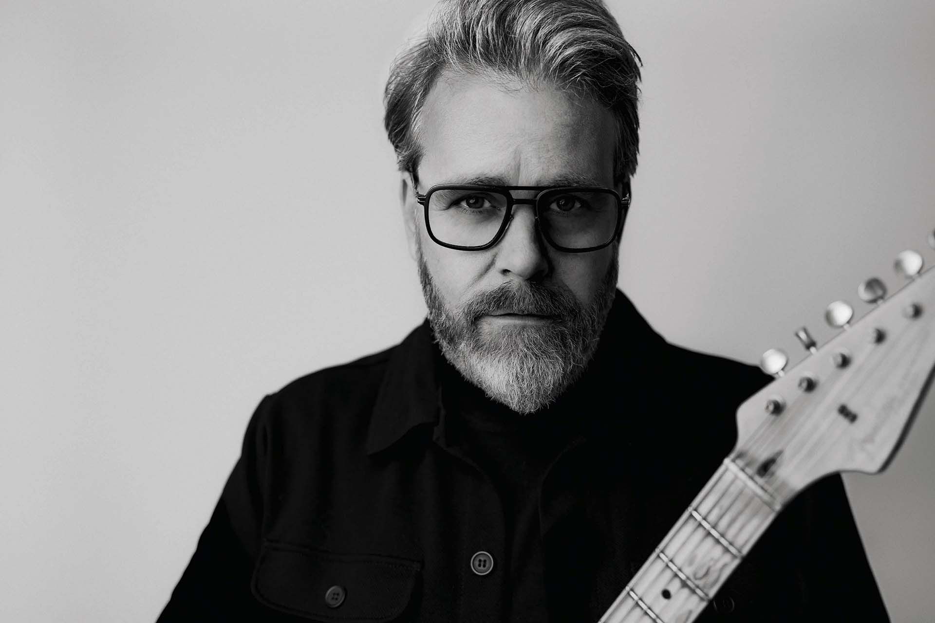 Zwart-wit portret van Ed Struijlaart met bril en baard die een elektrische gitaar vasthoudt en recht in de camera kijkt