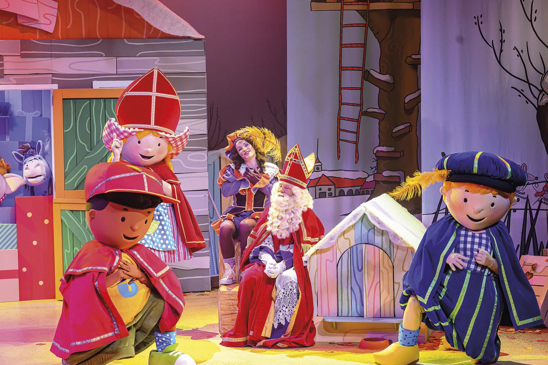 Sinterklaas en kleurrijke kinderfiguren dansen op een feestelijk podiumdecor met vlaggetjes en een huisje op de achtergrond