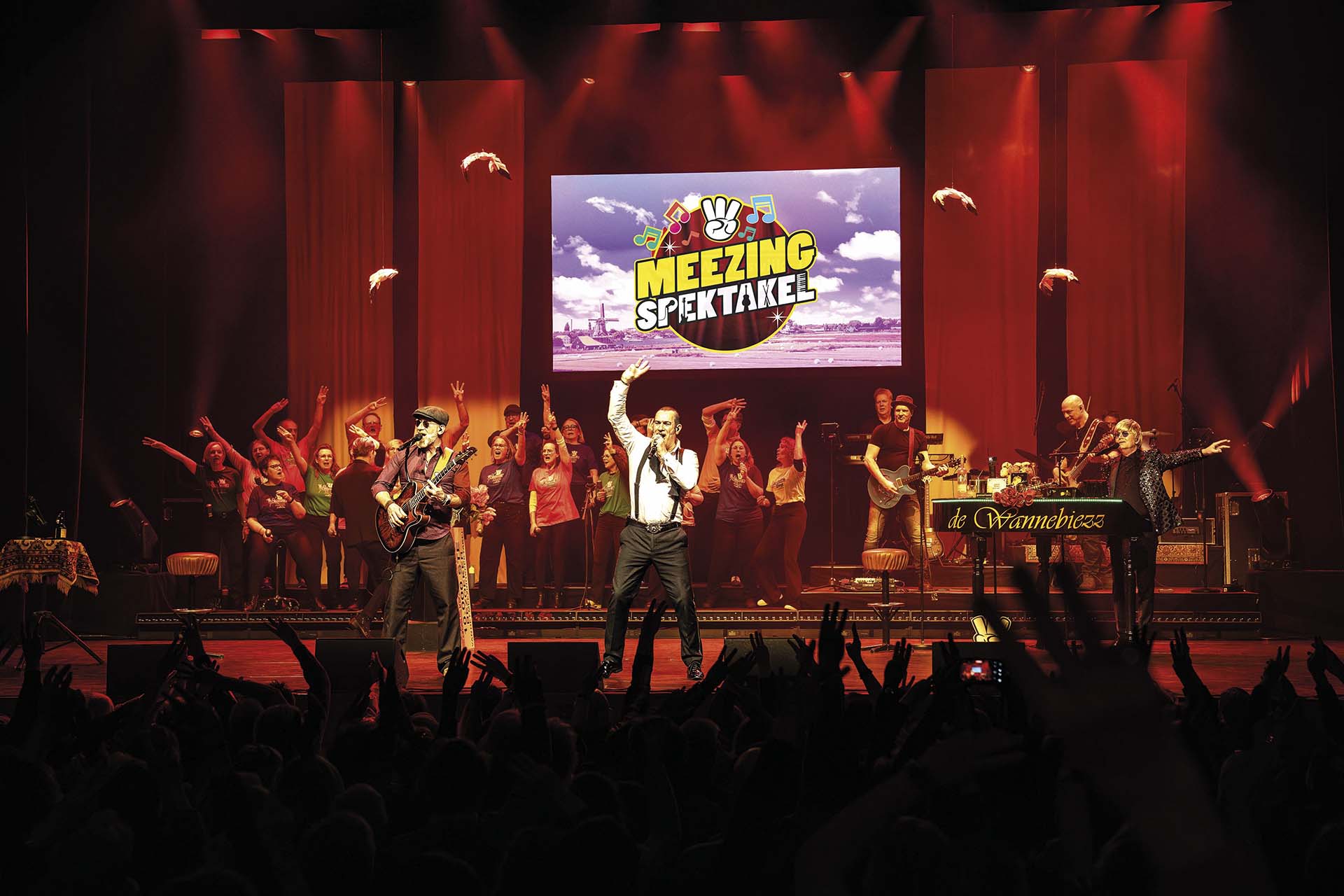 Een groep zangers en muzikanten staat op een verlicht podium met rode spotlights. Op de achtergrond zingt een koor, terwijl vooraan twee mannen zingen en gitaar spelen. Boven hen hangt een scherm met de tekst Meezing Spektakel. Het publiek steekt handen in de lucht.