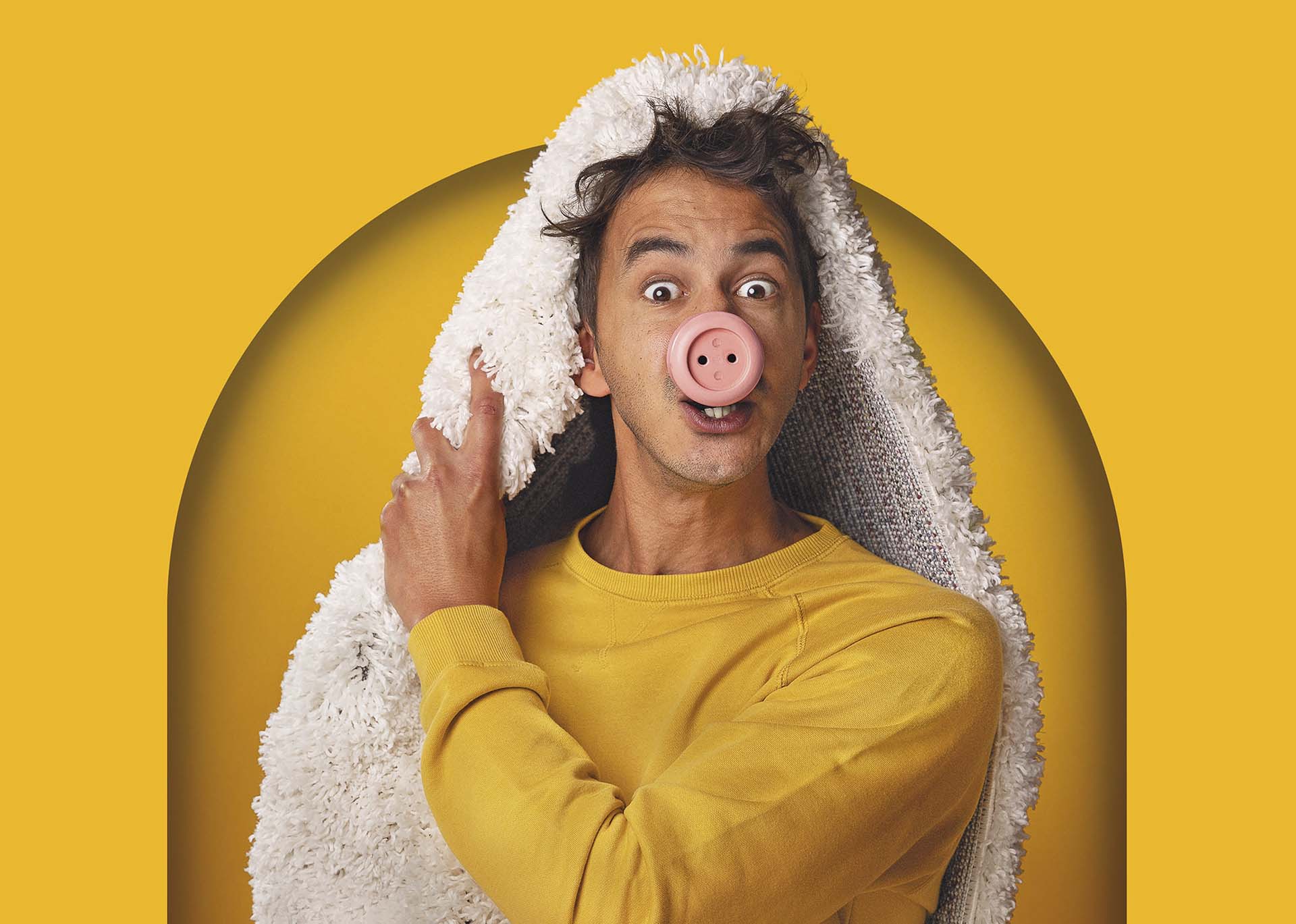 Man met speelse uitdrukking draagt een deken over zijn hoofd en een roze varkenssnuit, tegen felgele achtergrond in humoristische pose