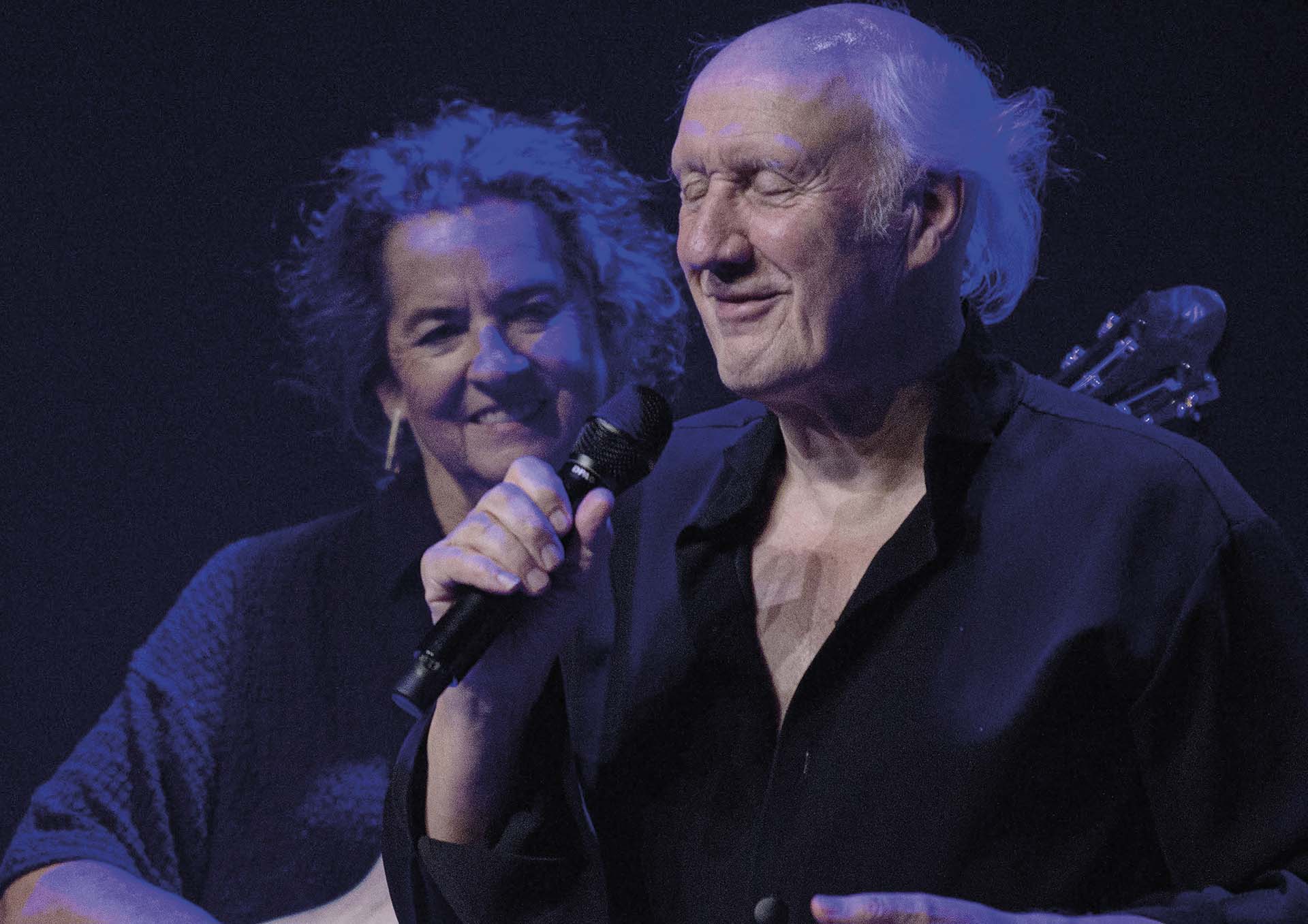 Herman van Veen staat op het podium met een microfoon in de hand, terwijl een muzikant achter hem glimlachend toekijkt.