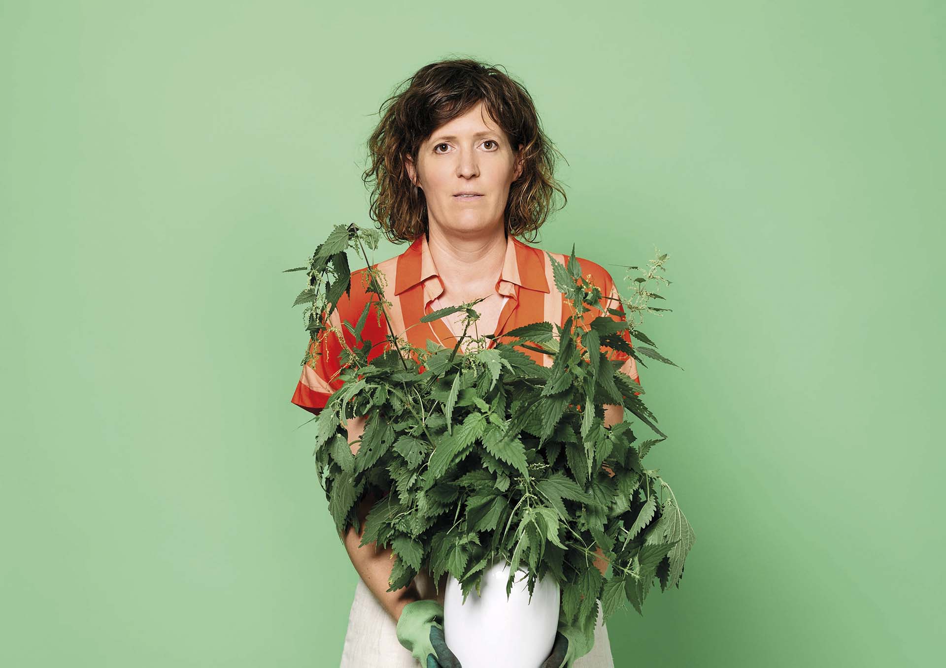 Janneke de Bijl in oranje blouse houdt grote bos brandnetels in witte vaas vast tegen groene achtergrond, kijkt recht in camera met neutrale uitdrukking