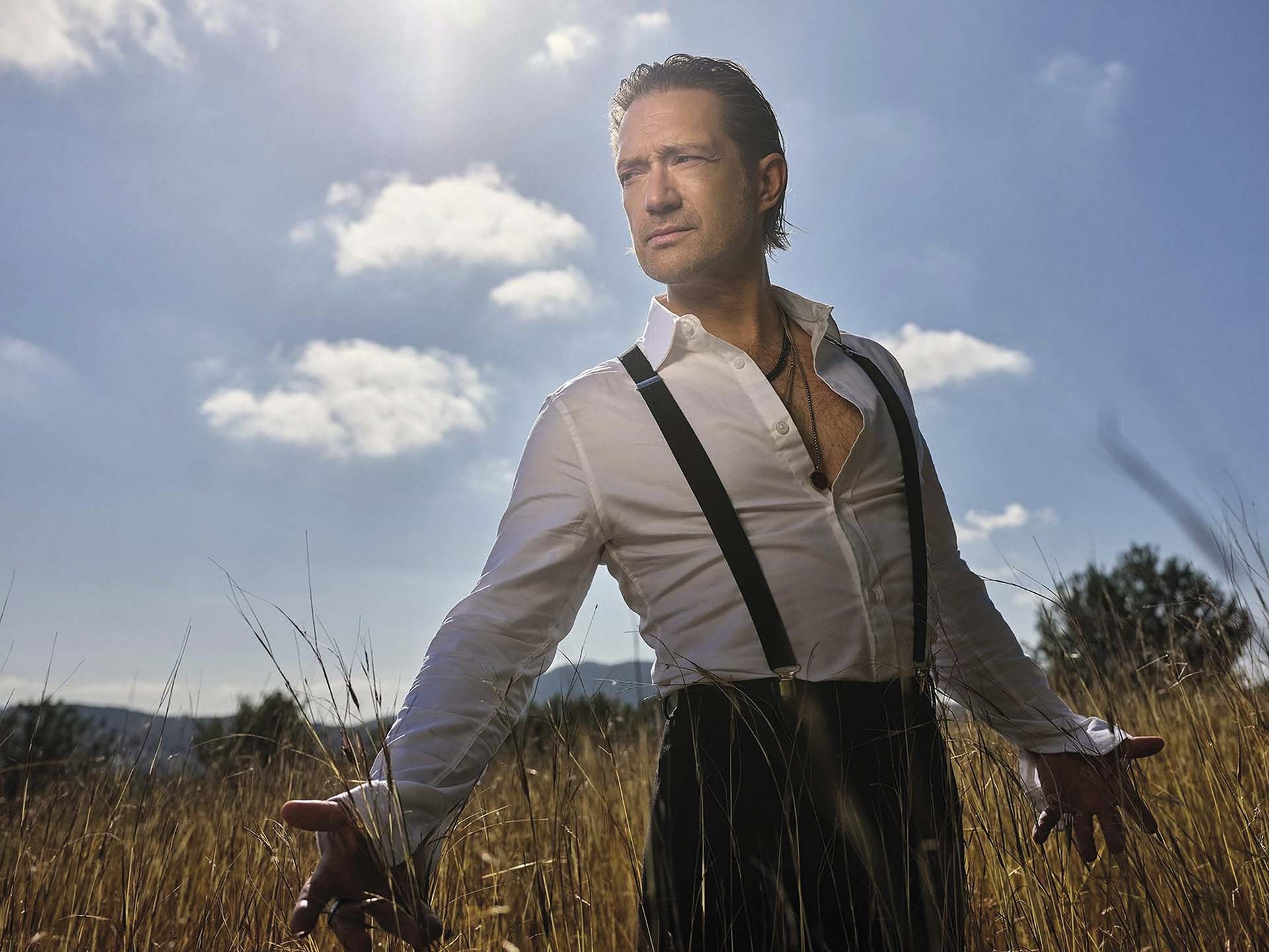Joost Spijkers in wit overhemd met bretels staat in een veld met hoog gras en kijkt in de verte onder een zonnige blauwe lucht