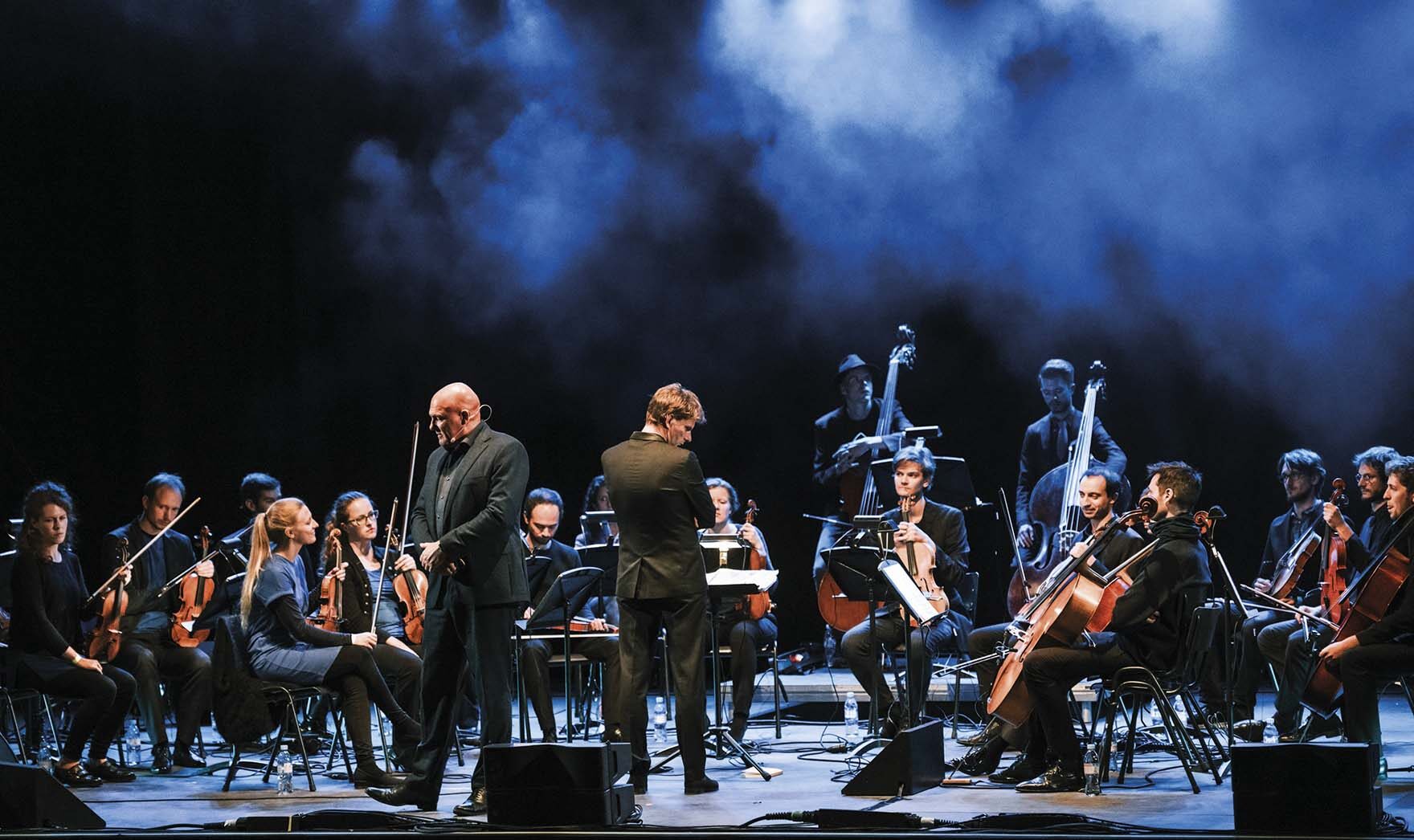 Orkest met strijkers zit op podium terwijl dirigent en Andre Kuipers centraal staan, omgeven door blauwe belichting en rook tijdens een concertuitvoering