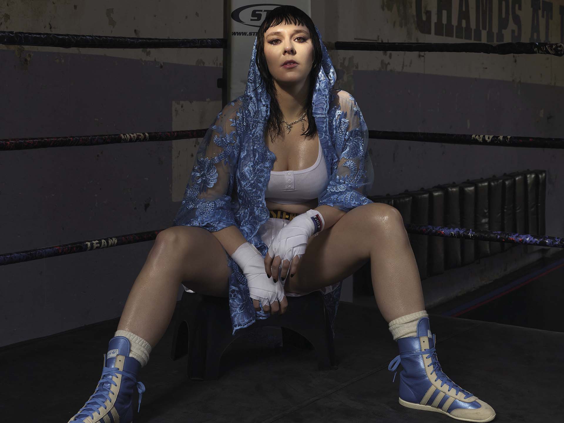 Kim Karssen zit in een boksring op een kruk met bokshandschoenen en blauwe jas, kijkt recht in de camera met een krachtige houding