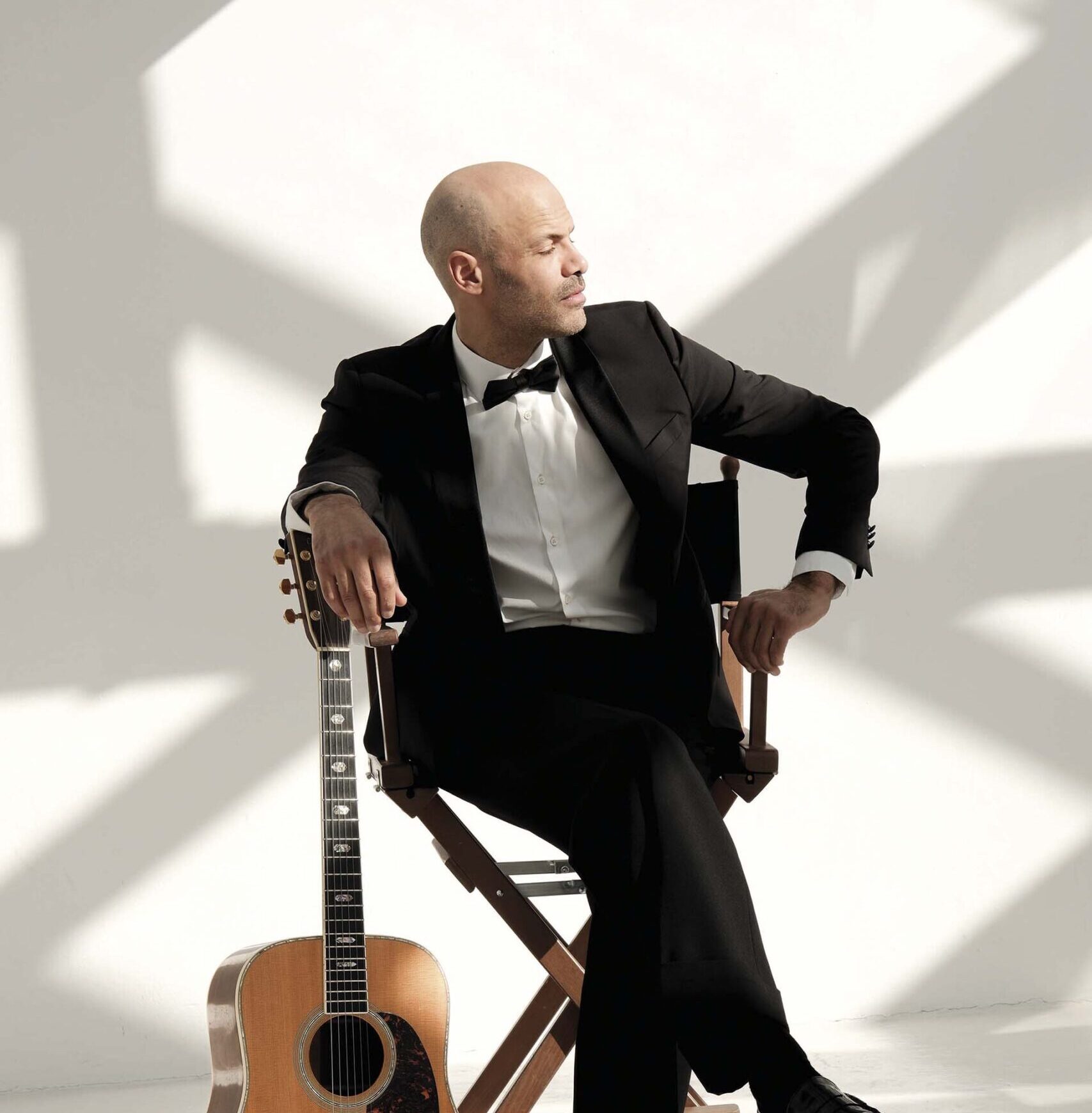Alain Clark zittend in een stoel. Hij heeft een mooi pak aan en leunt met zijn arm op een gitaar.